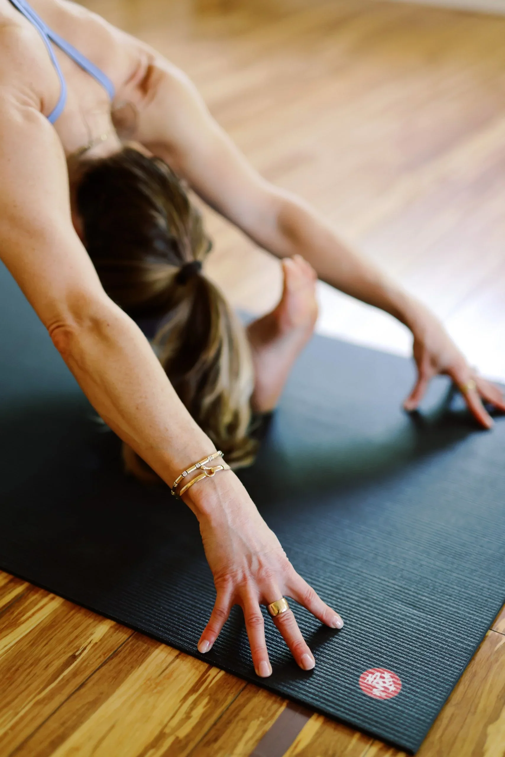 A person practicing yoga on a black mat, in a downward dog pose with hands and feet on the mat, wearing a light blue sports bra, with a wooden floor and a red and white circular logo on the mat visible.