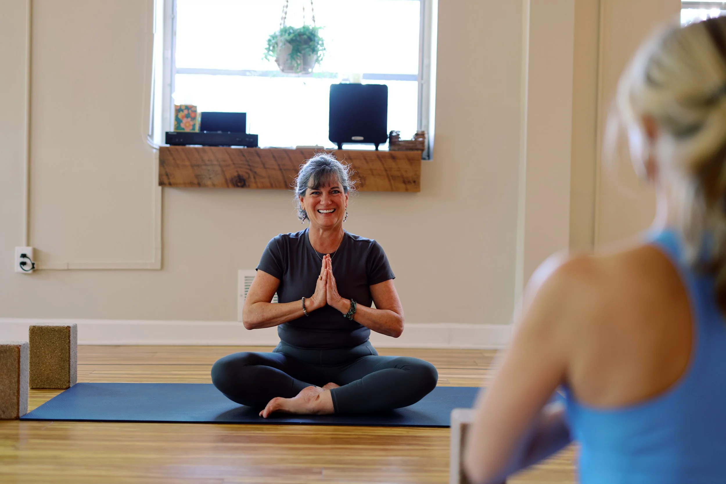 A woman sitting on a yoga mat in a seated twist pose, smiling with hands in a prayer position, facing a child in yoga attire in a yoga studio with wooden flooring and a window.