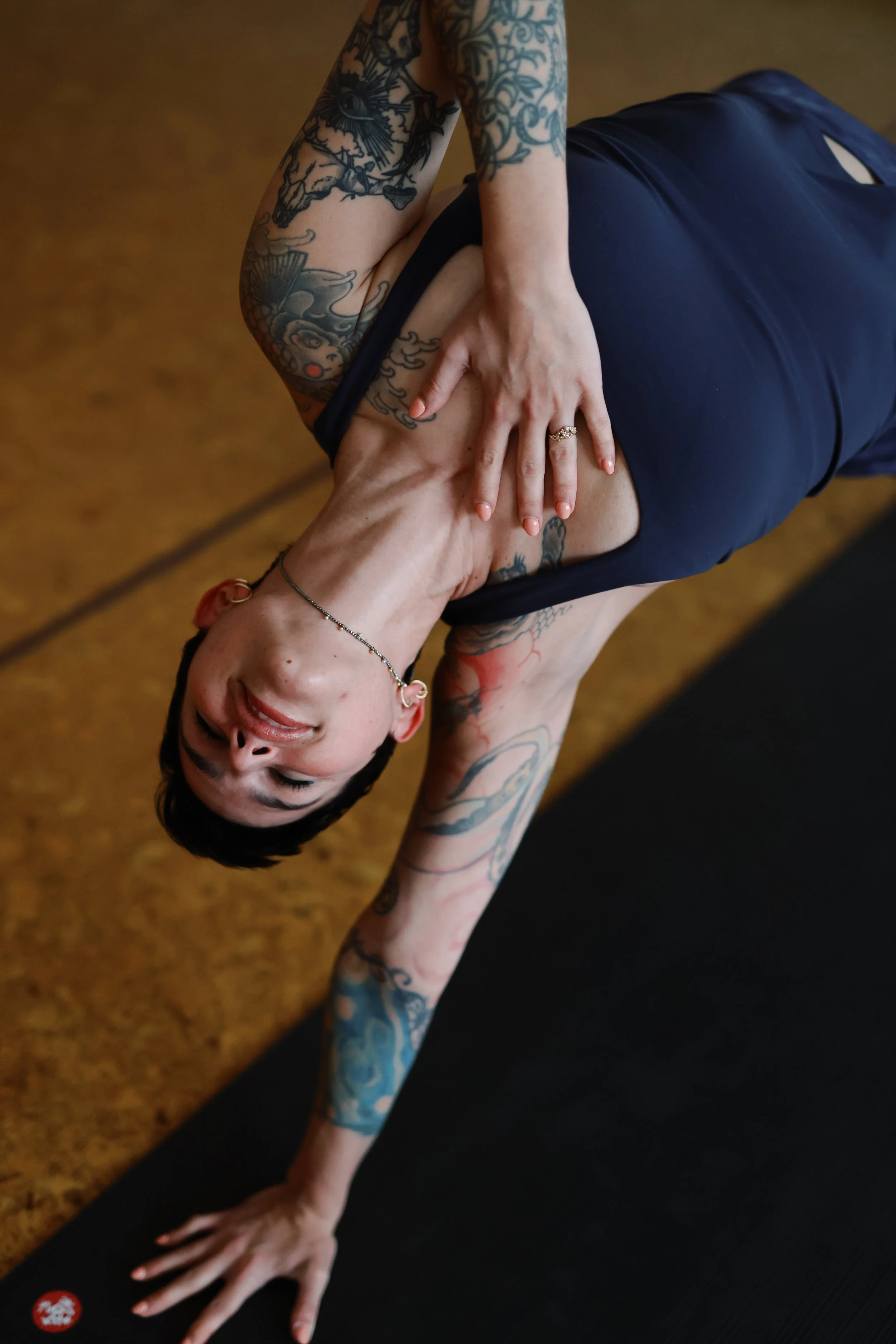 A woman with tattoos performing a yoga pose on a black mat, wearing a blue sports bra and matching shorts, in a room with a wooden floor.