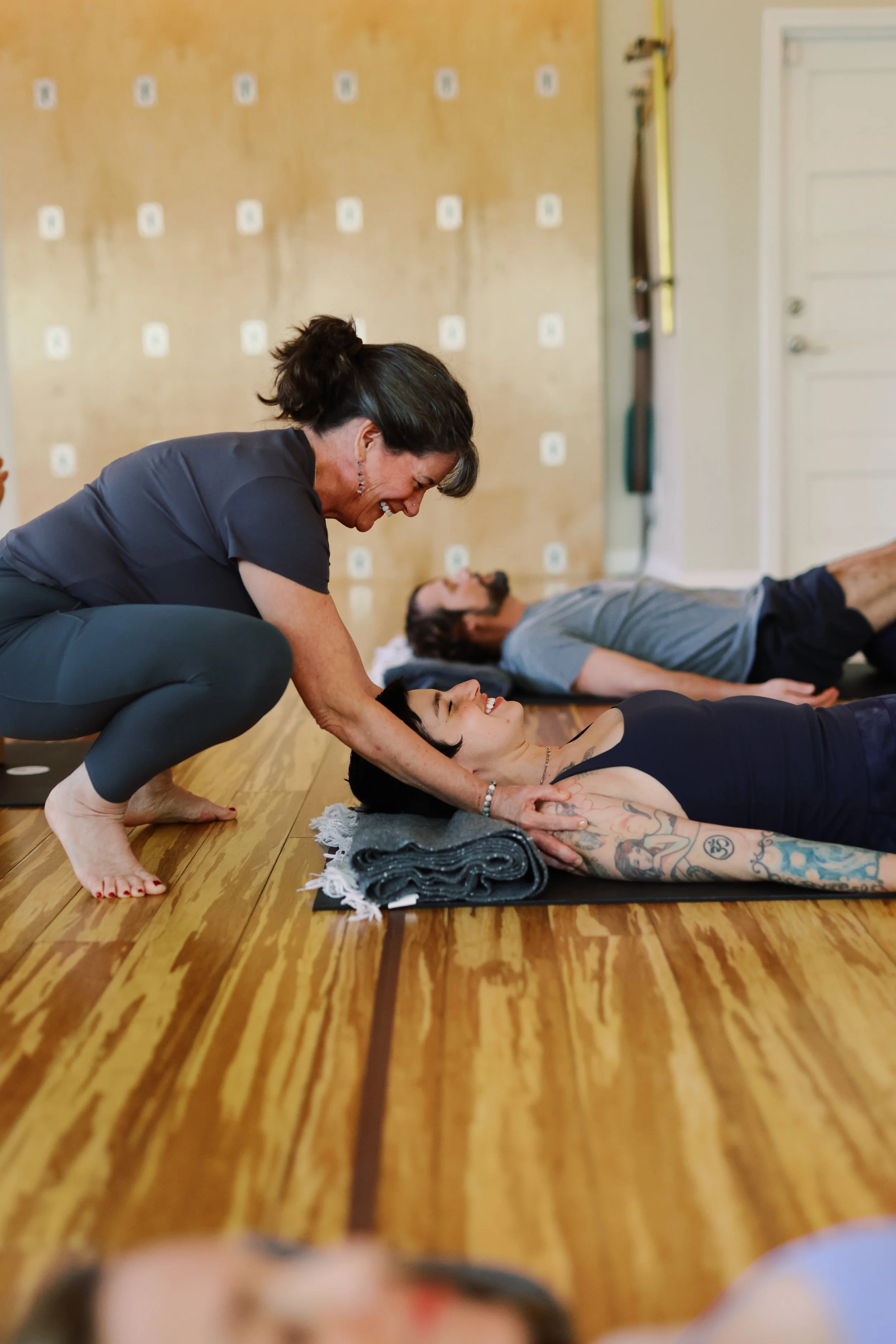 A woman practicing yoga on a mat with a rolled-up towel beside her, while an instructor assists her in a supported backbend pose in a yoga studio. Two other people are practicing in the background.