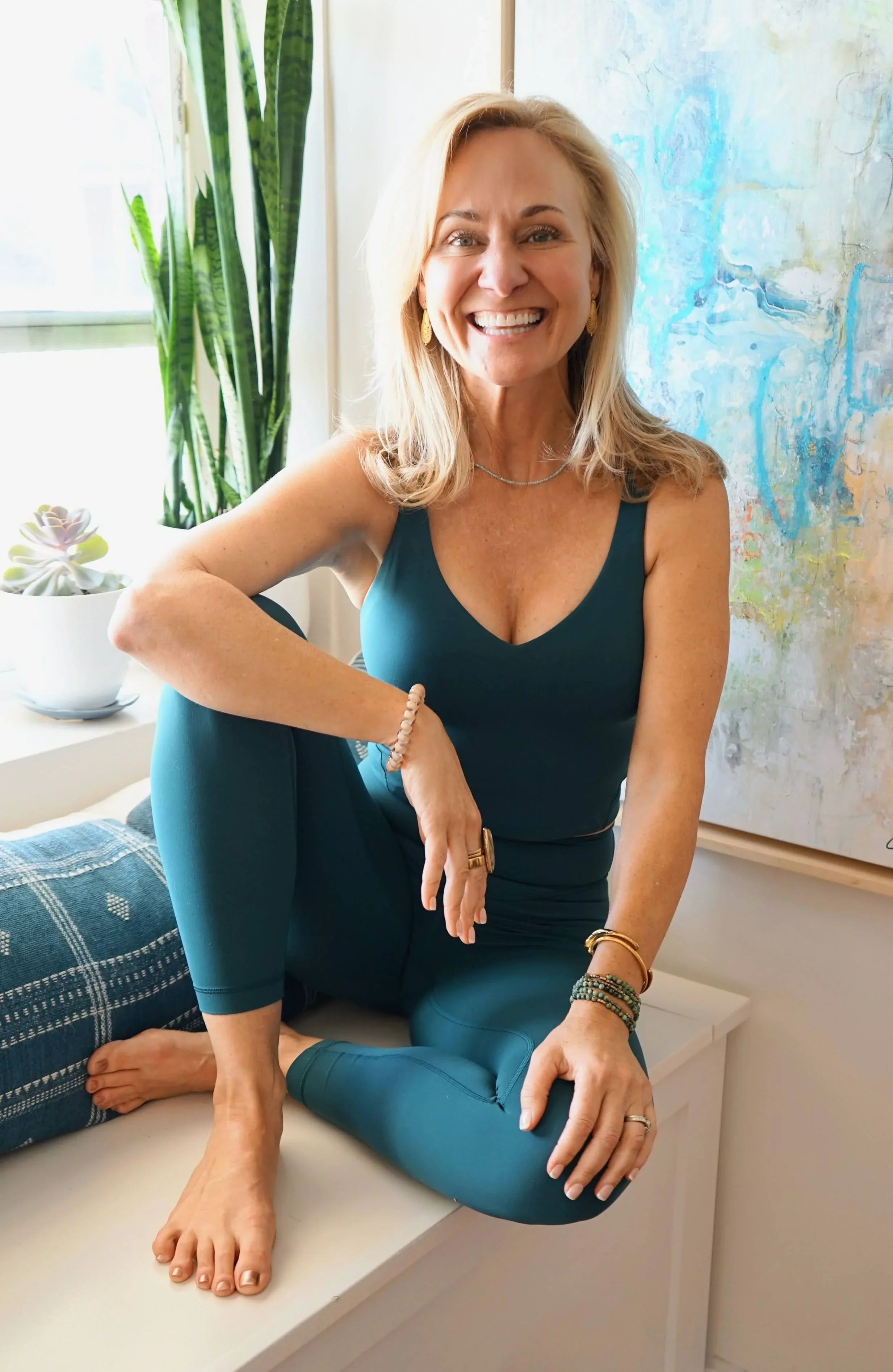 A woman with blonde hair smiling and sitting on a bed, while another person with light skin is performing a yoga pose with their foot on her shoulder. The person on the bed is wearing a teal tank top and teal leggings, and the other person's foot is bare. There is a green plant and an abstract painting in the background.