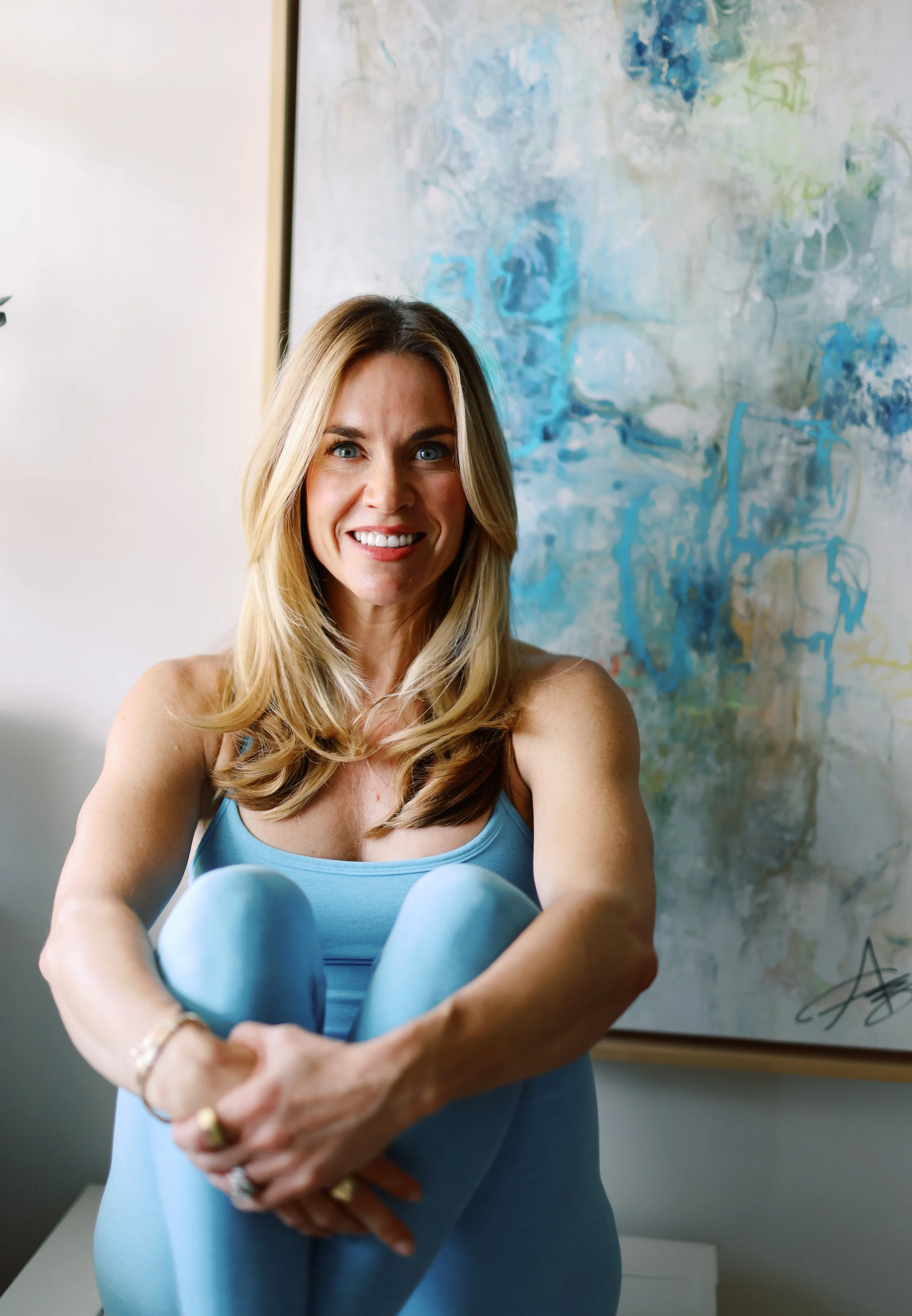 A woman with long blonde hair and blue eyes taking a selfie, wearing a blue tank top, with an abstract painting in shades of blue and green in the background.
