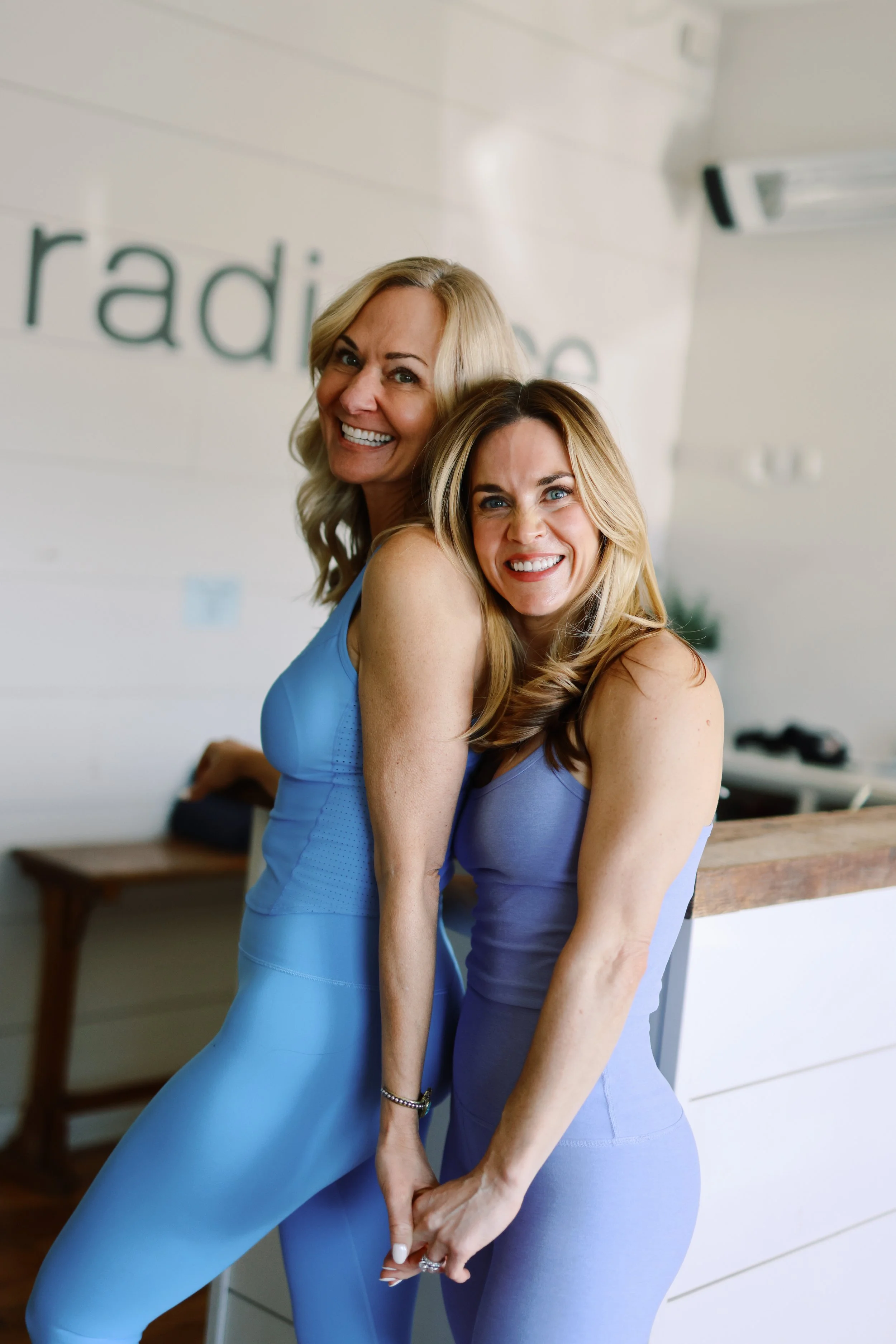 Two women in blue workout clothing smiling and holding hands in a bright, modern room with white walls and wooden furniture.
