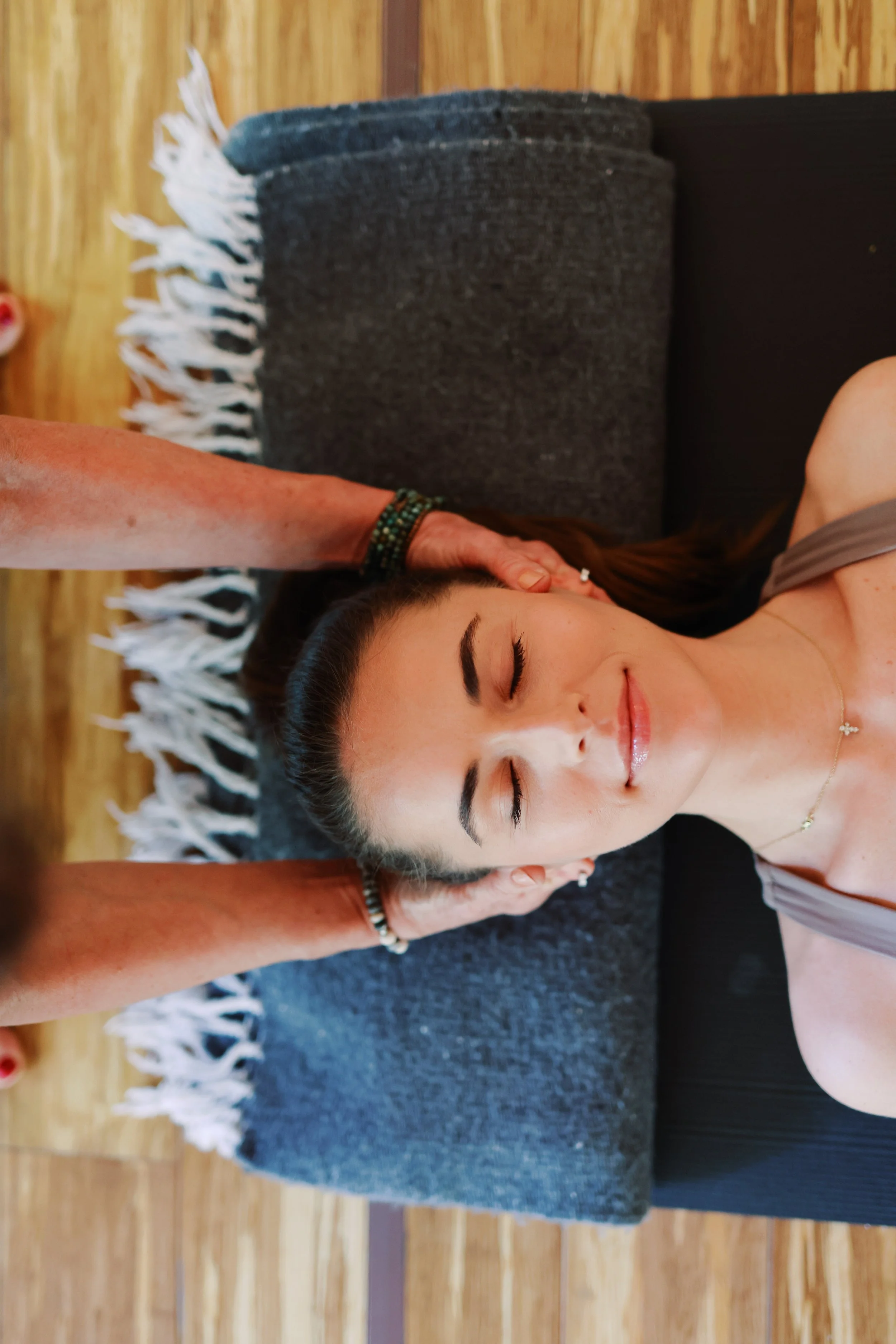 A young woman with dark hair, closed eyes, and a gentle smile lying on her side on a yoga mat, receiving a head massage from a person with a beaded bracelet, in a room with wooden flooring and a woven fabric blanket or rug in the background.