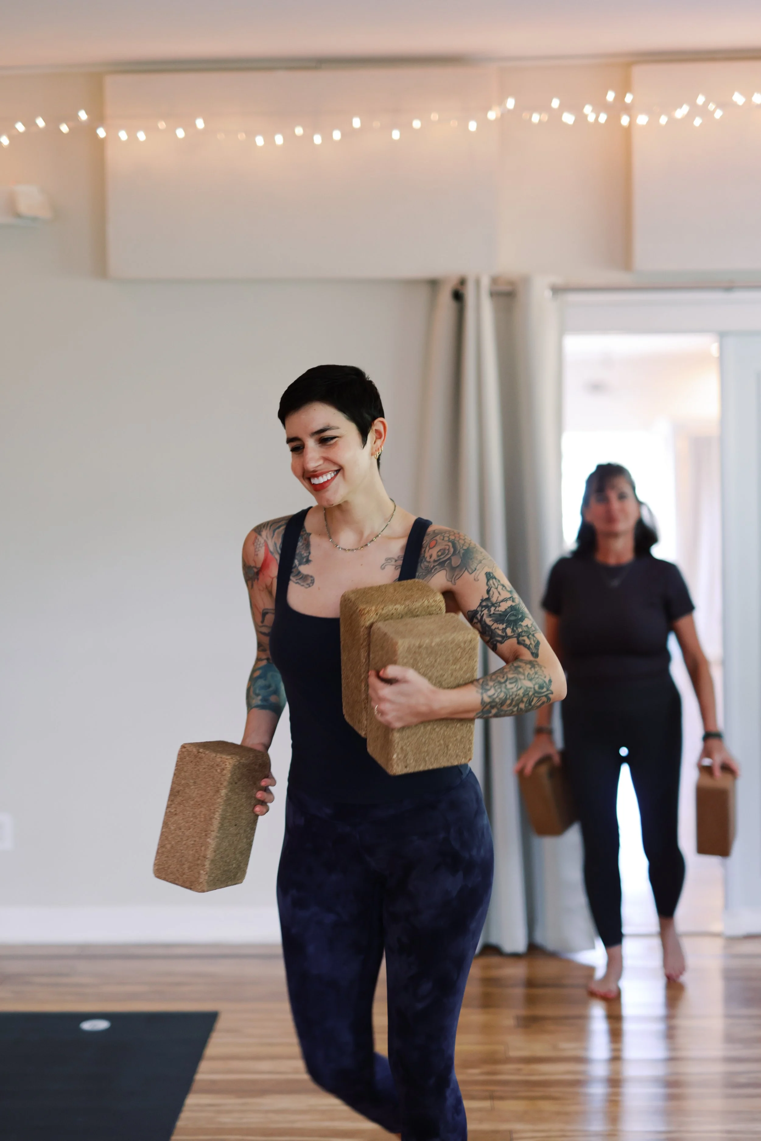 A woman with short dark hair and tattoos on her arms smiling as she holds a yoga block in a room with wooden flooring, a yoga mat, and string lights on the ceiling. In the background, another woman with dark hair, wearing black, is also holding yoga blocks.