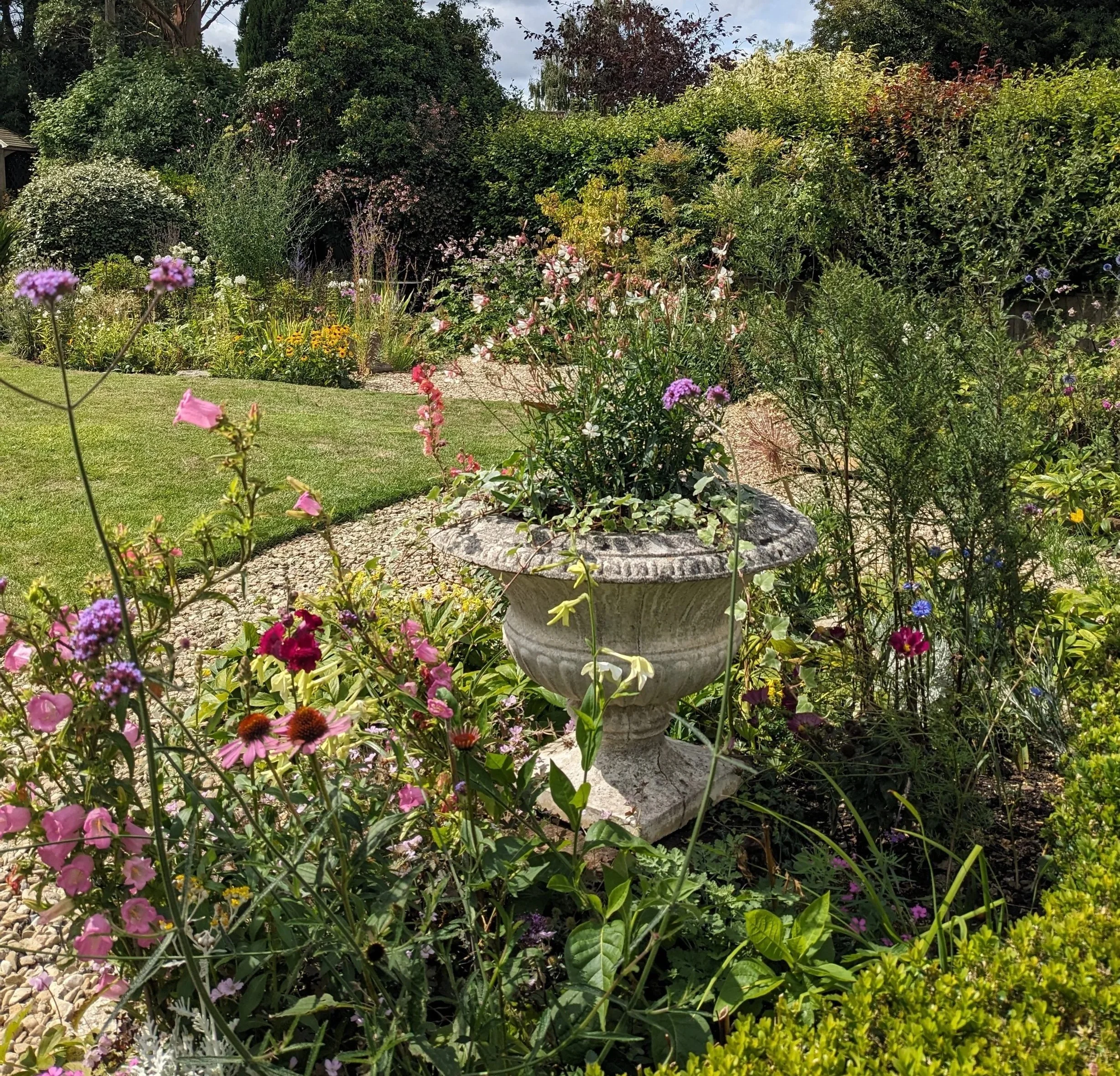 A lush garden with a variety of colorful flowers, green bushes, and trees. There is a stone birdbath in the center among the flowers, and a well-maintained lawn surrounds the garden. Cloudy sky can be seen in the background.