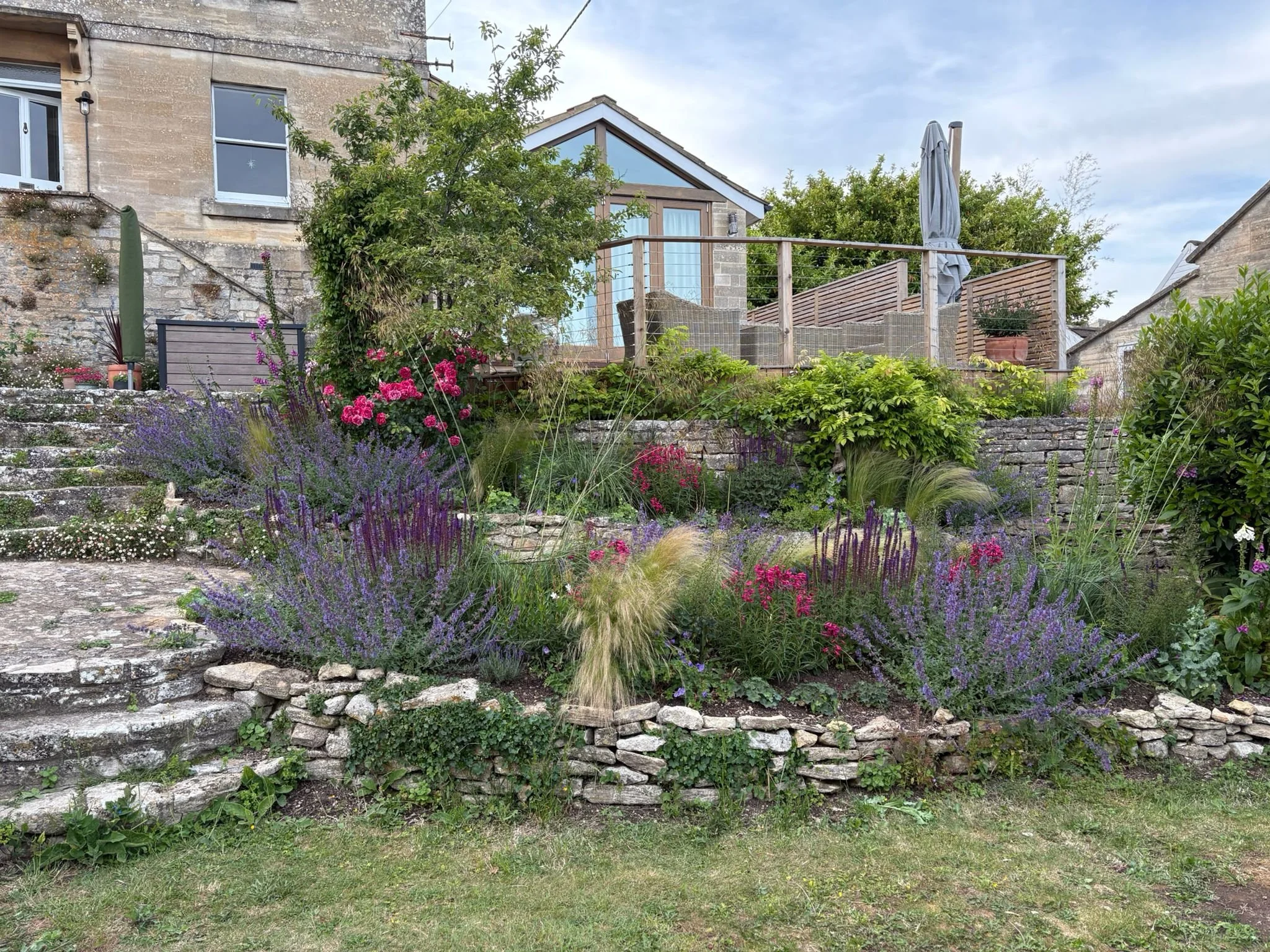 A garden with colorful flowers and plants, stone steps, and a house with a balcony in the background.