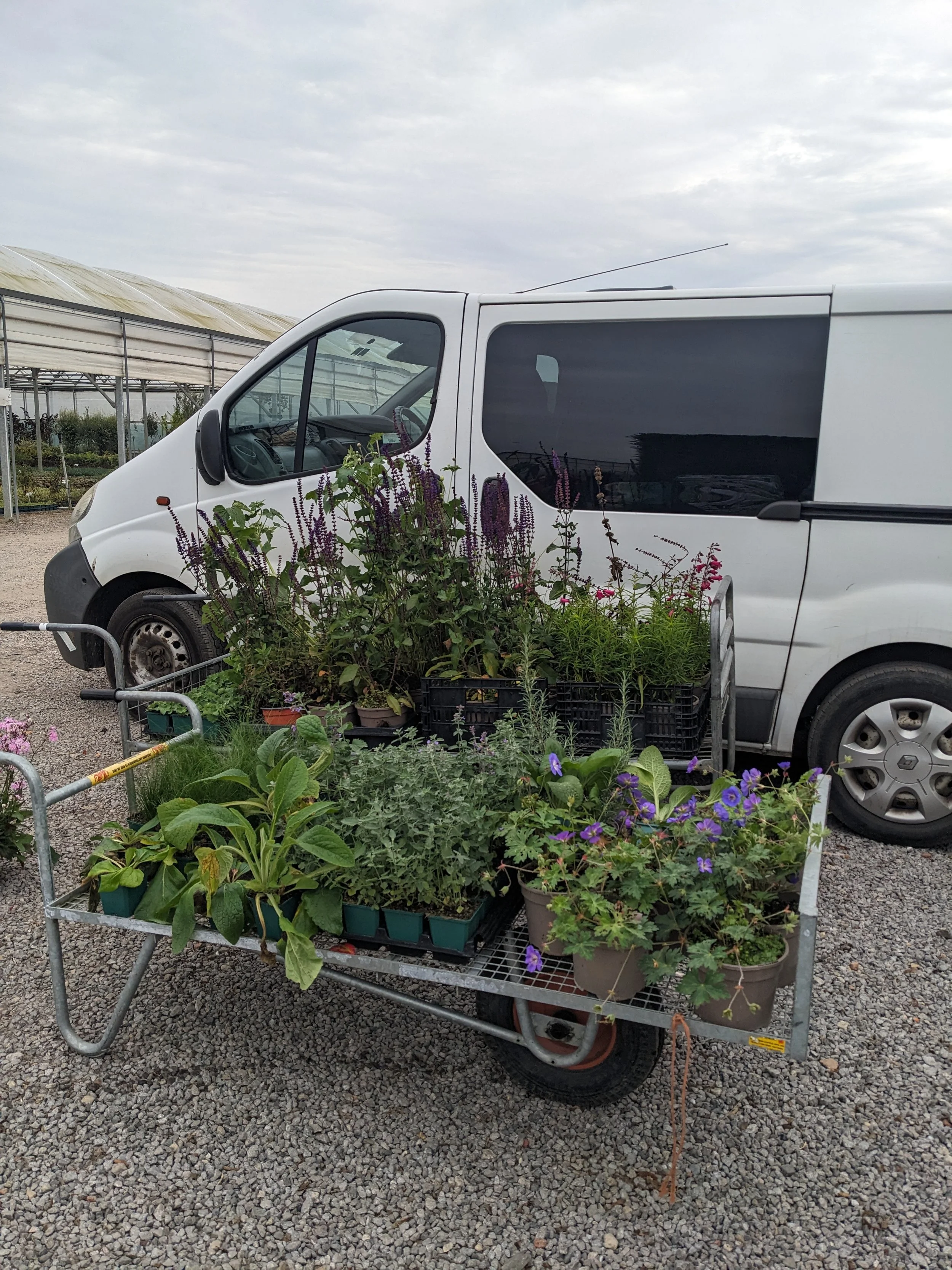 A white van parked outdoors with a large basket of potted plants and flowers in front of it, near a greenhouse and gravel ground.