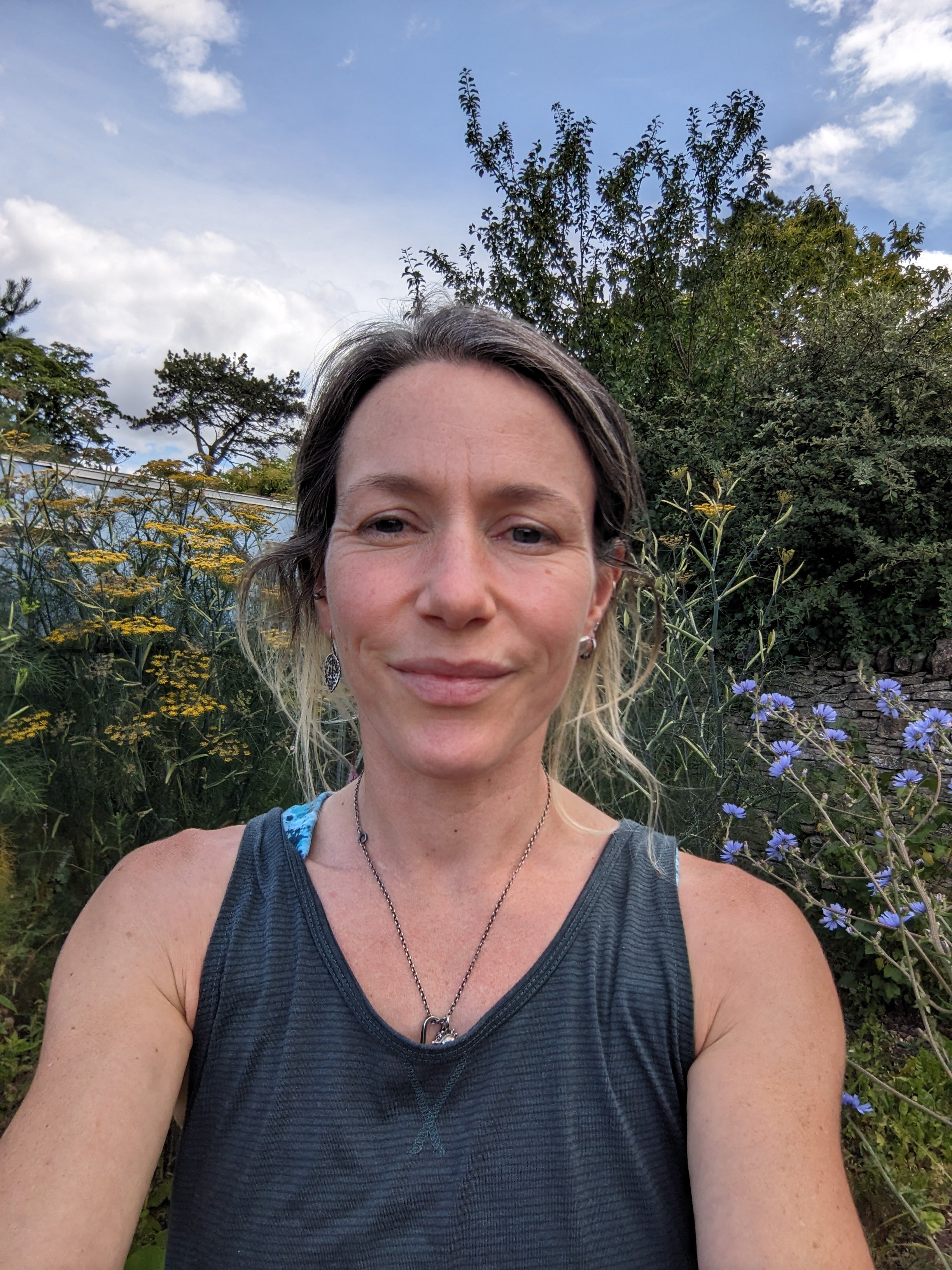 A woman with shoulder-length light brown hair taking a selfie outdoors, with greenery and flowering plants behind her, under a partly cloudy sky.