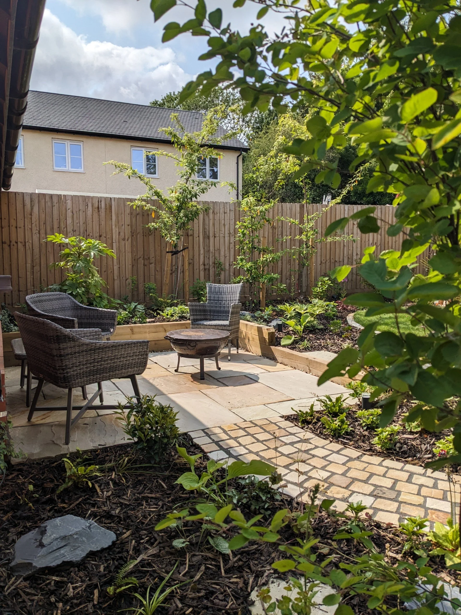 A backyard garden with a patio area featuring chairs and a small fire pit, surrounded by various plants and trees, with a wooden fence and a house with white windows in the background.
