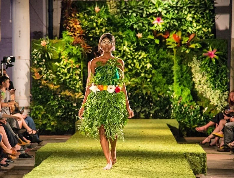 Model walking down a moss runway, wearing a dress made of plants and flowers, with lush living walls as backdrop.