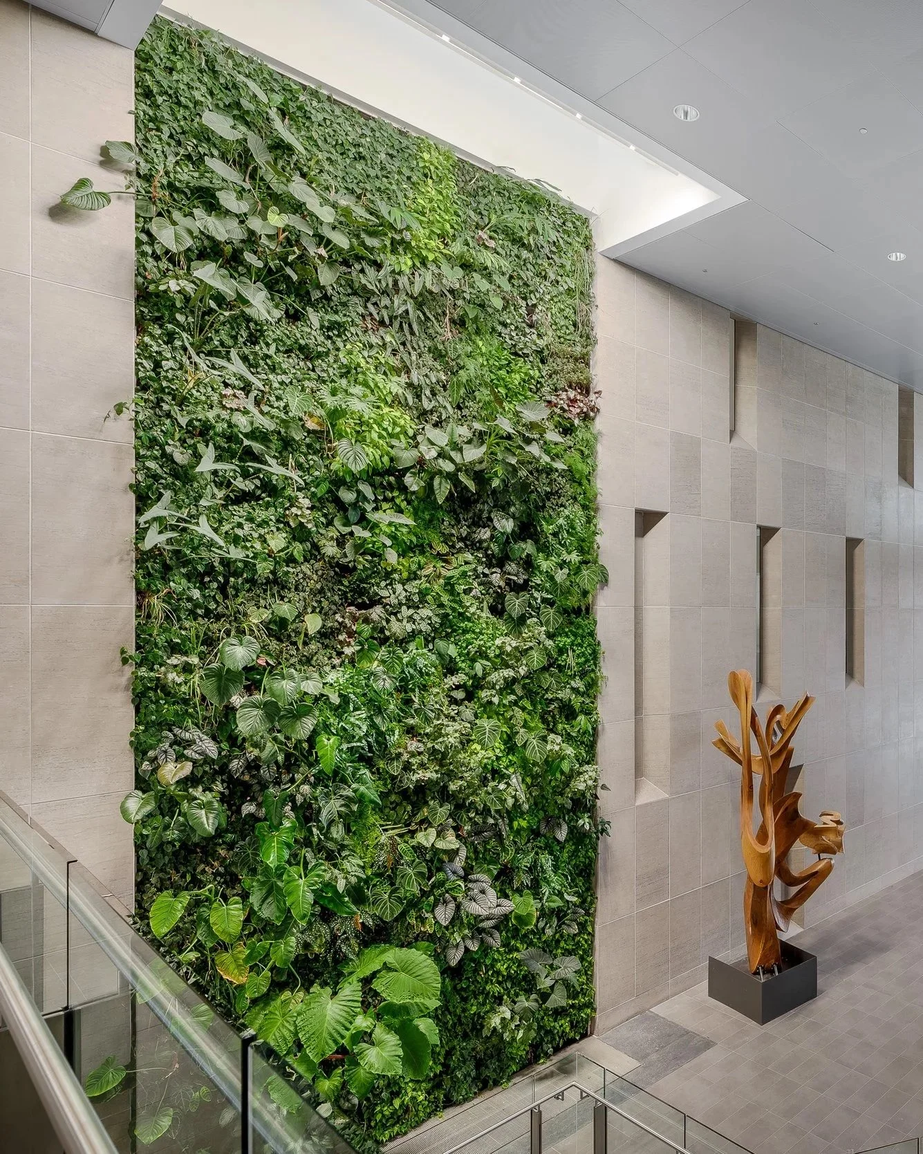 Indoor vertical garden with various green plants and large leaves on a wall, with a wooden abstract sculpture nearby.