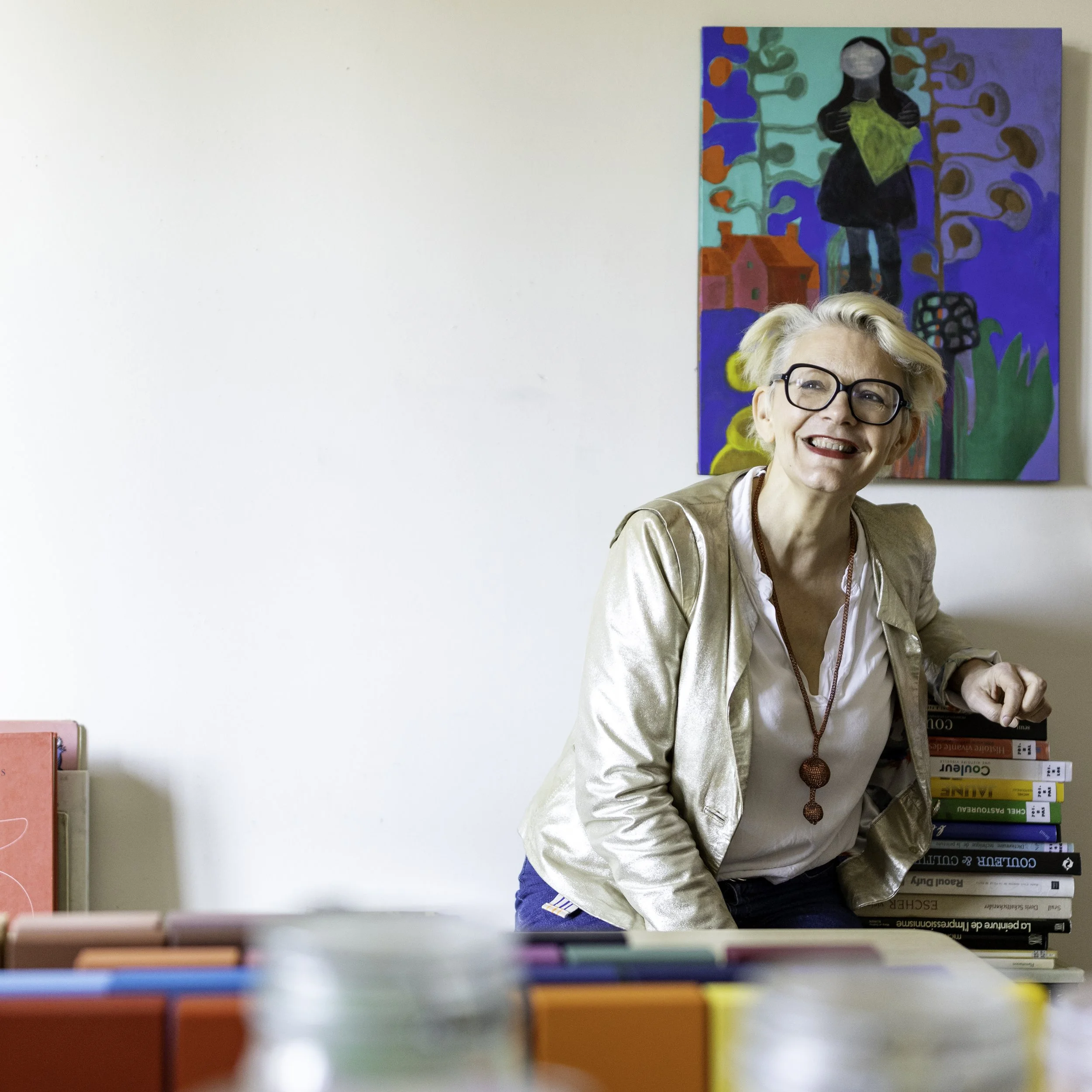 Une femme âgée souriante avec des lunettes noire, portant une veste beige et une chaîne enroulée autour de son cou, se tient à côté d'une pile de livres, dans une pièce bien éclairée, avec une peinture artistique colorée accrochée au mur derrière elle.
