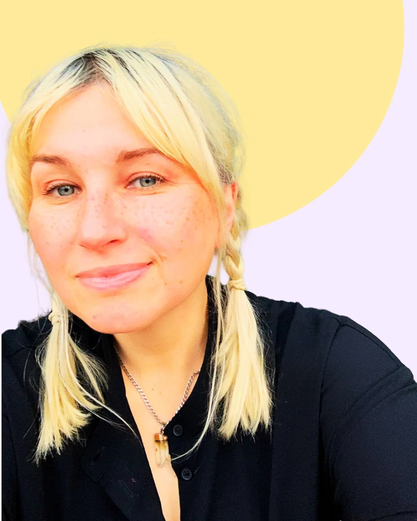 Close-up portrait of a young woman with blonde hair in braids, blue eyes, and freckles, smiling gently, wearing a black shirt and a necklace with a pendant. The background features a pastel purple and yellow design.