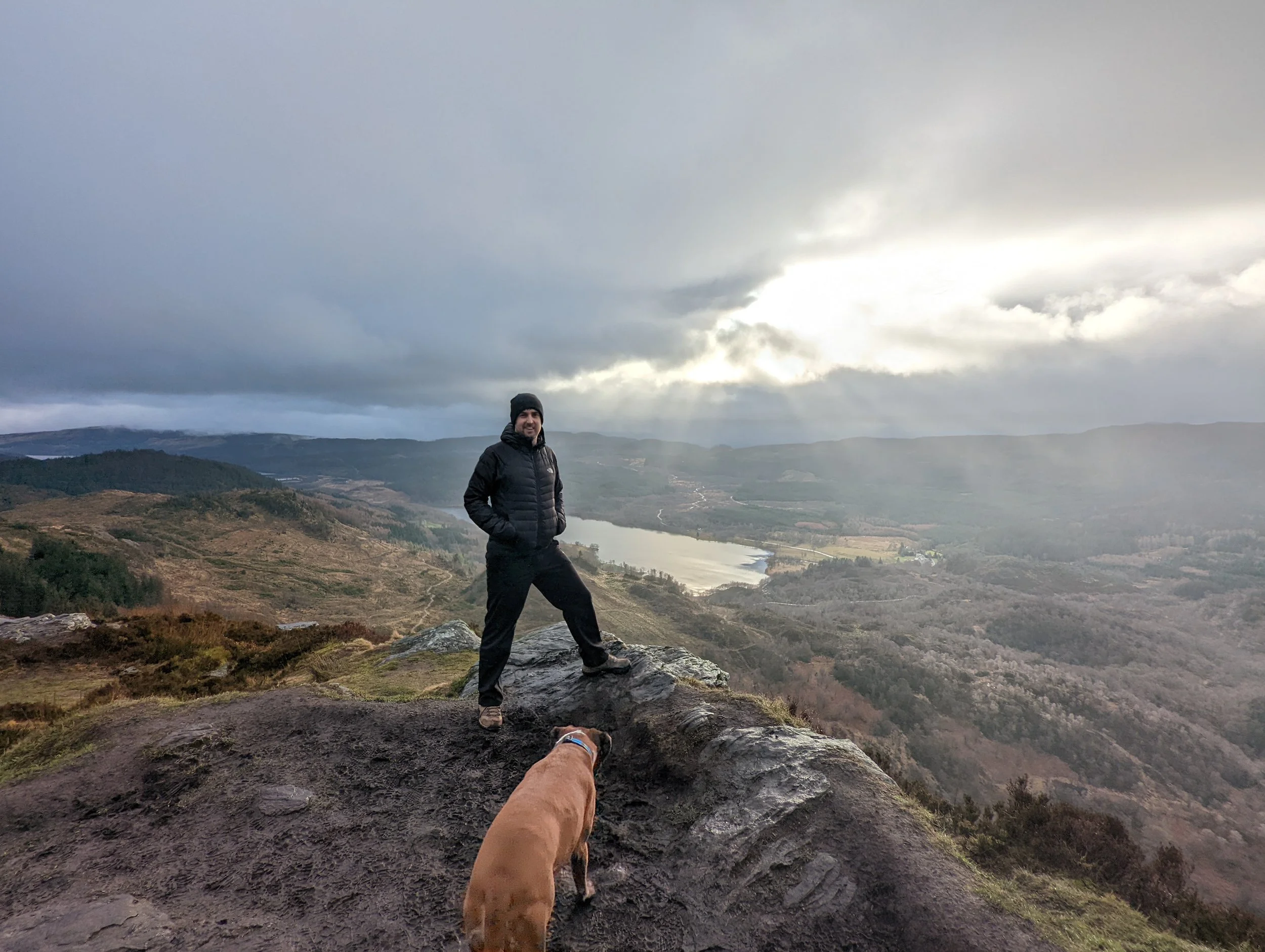 A man in black outdoor clothing standing on a large rock at a mountain overlook with scenic landscape, river, and cloudy sky in the background, with a brown dog nearby.