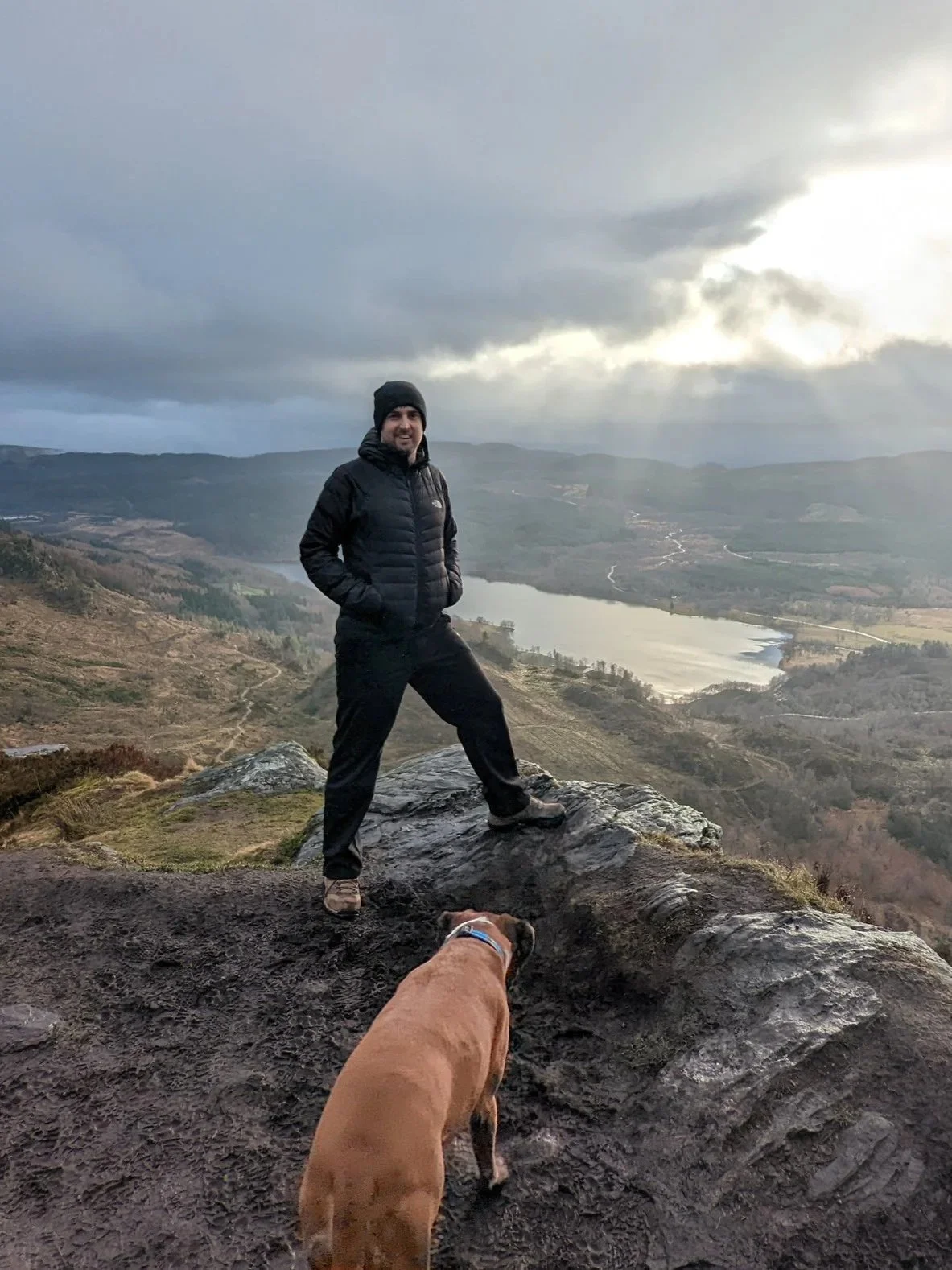 A man standing on a rock at a high vantage point overlooking a valley with a river and forested hills, wearing a black jacket and beanie, with a brown dog in the foreground.