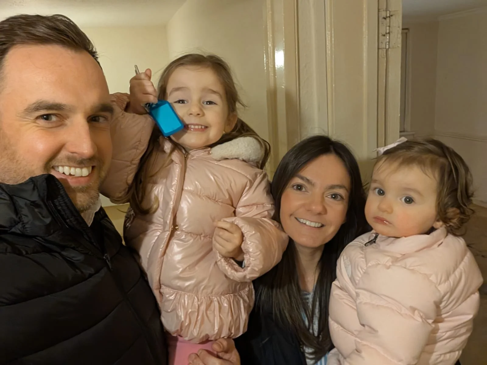 A family of four taking a selfie indoors, with a man and woman smiling, and two young girls in light pink jackets, one holding a toy walkie-talkie.