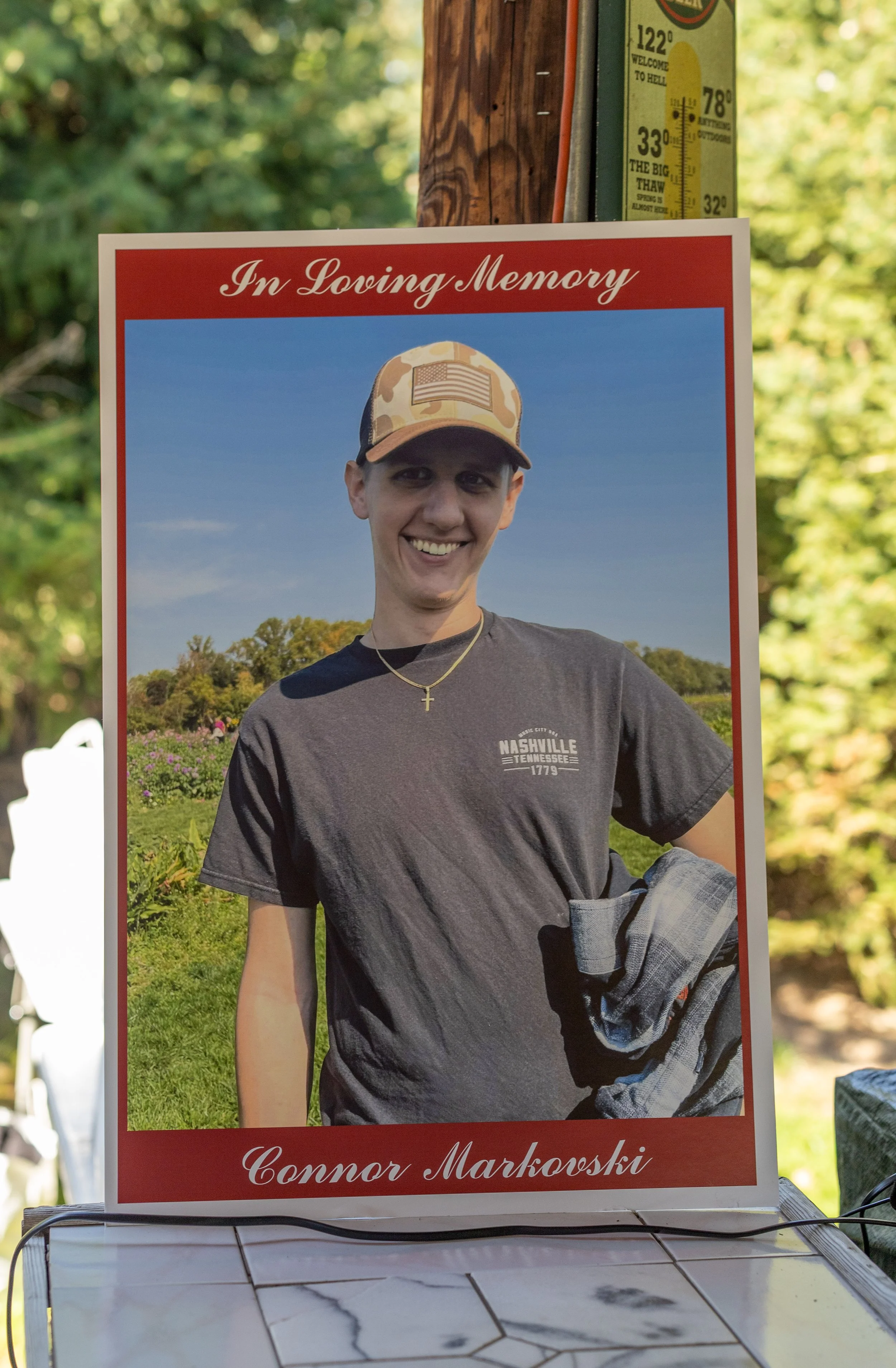 A memorial sign with a photo of a smiling young man wearing a camouflage cap with an American flag patch, a cross necklace, and a Nashville Tennessee t-shirt, set outdoors with trees and a blue sky in the background. The top of the sign says "In Loving Memory" and the bottom has the name "Connor Markowski."
