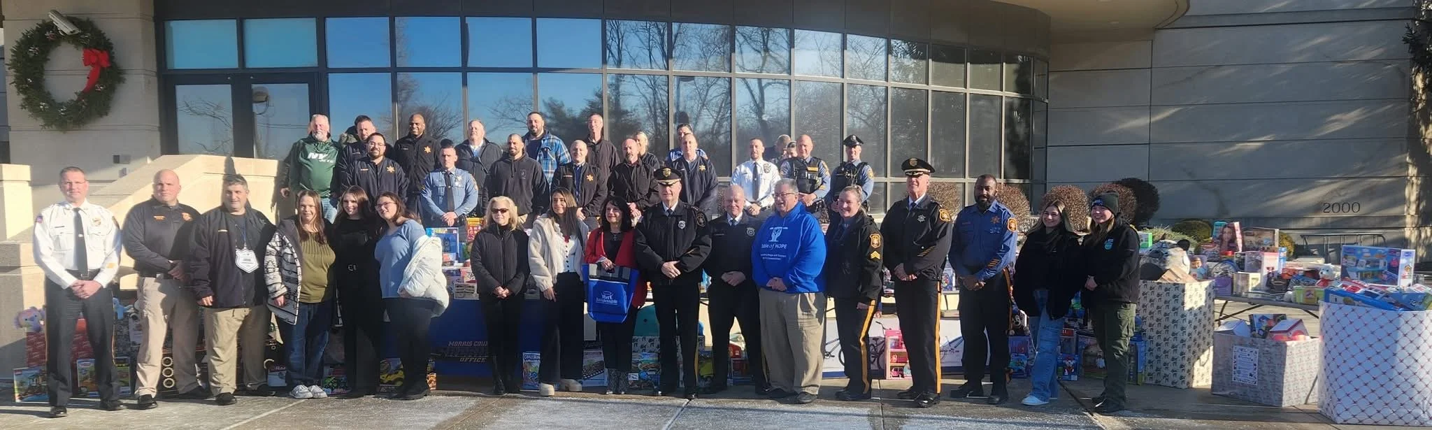 Group of police officers, community members, and volunteers standing outside a building with donation boxes for a charity event, decorated with a Christmas wreath.