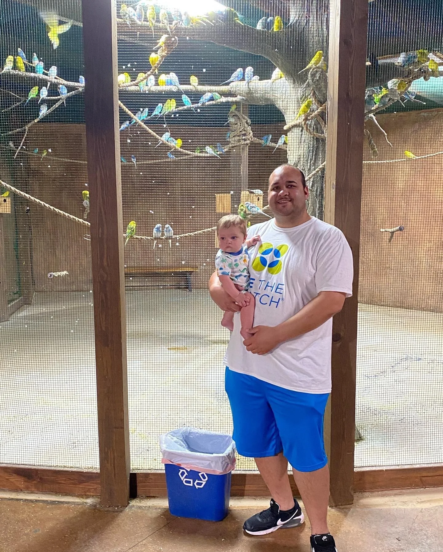 A man holding a young child stands in front of a bird cage filled with colorful birds on branches.