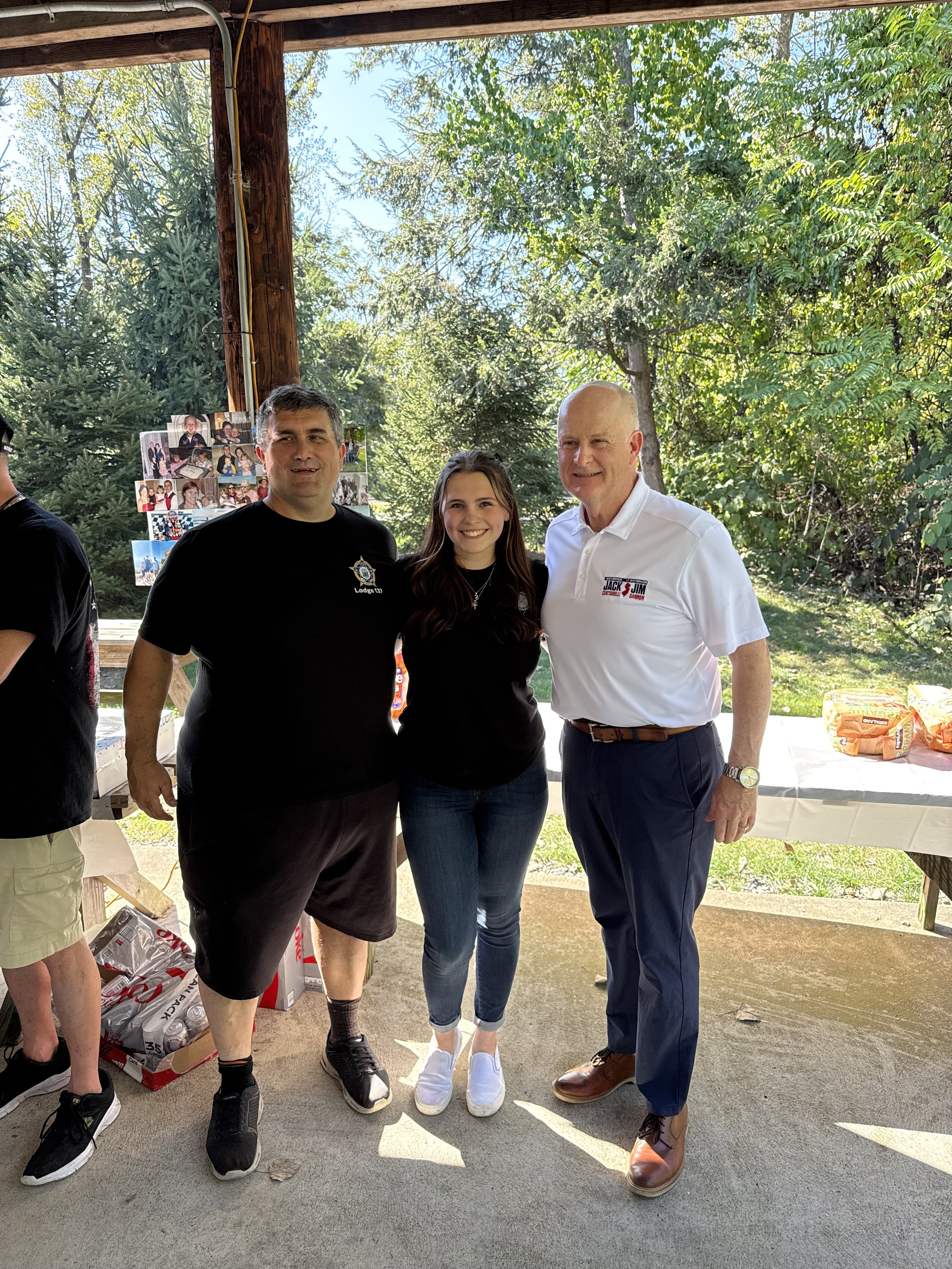 Three people standing outdoors under a wooden shelter, with trees in the background. The person on the left is a man in a black T-shirt and shorts, the person in the middle is a young woman in jeans and a black shirt, and the person on the right is a