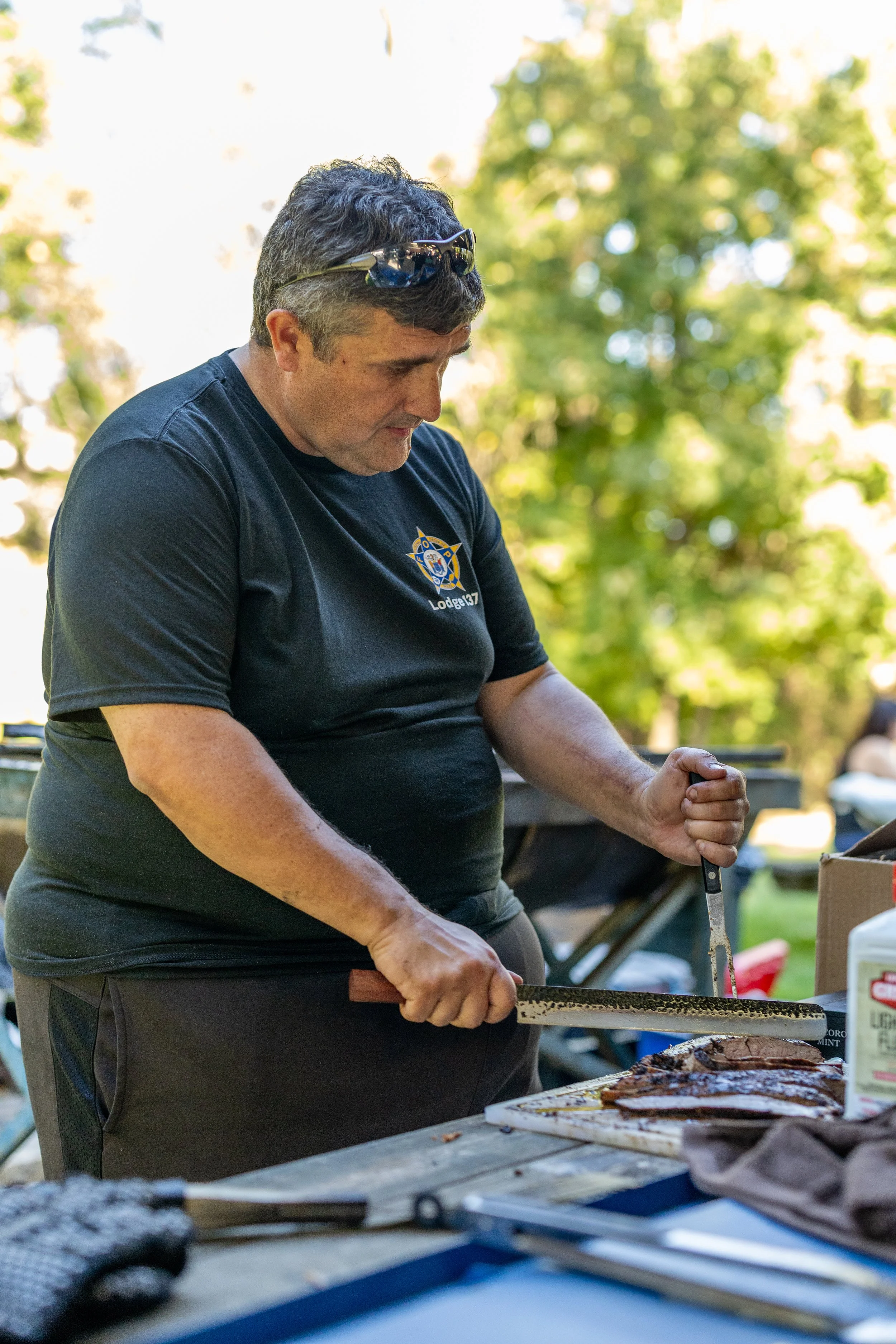 A man in a black shirt with a police badge emblem cuts meat with a knife on a wooden table outdoors in a park with trees in the background.