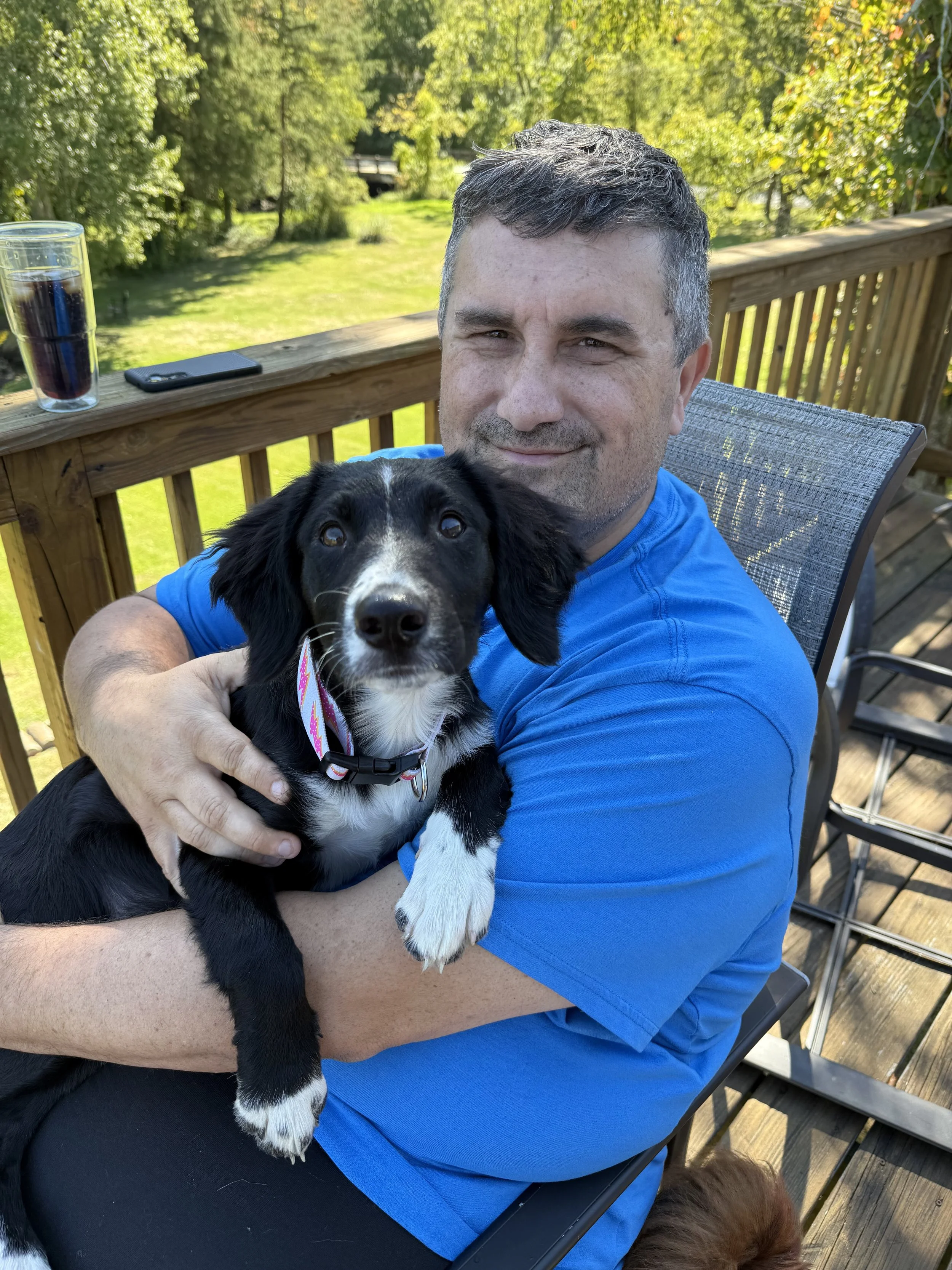 Man in blue shirt sitting on a wooden deck holding a black and white puppy, with greenery and trees in the background.