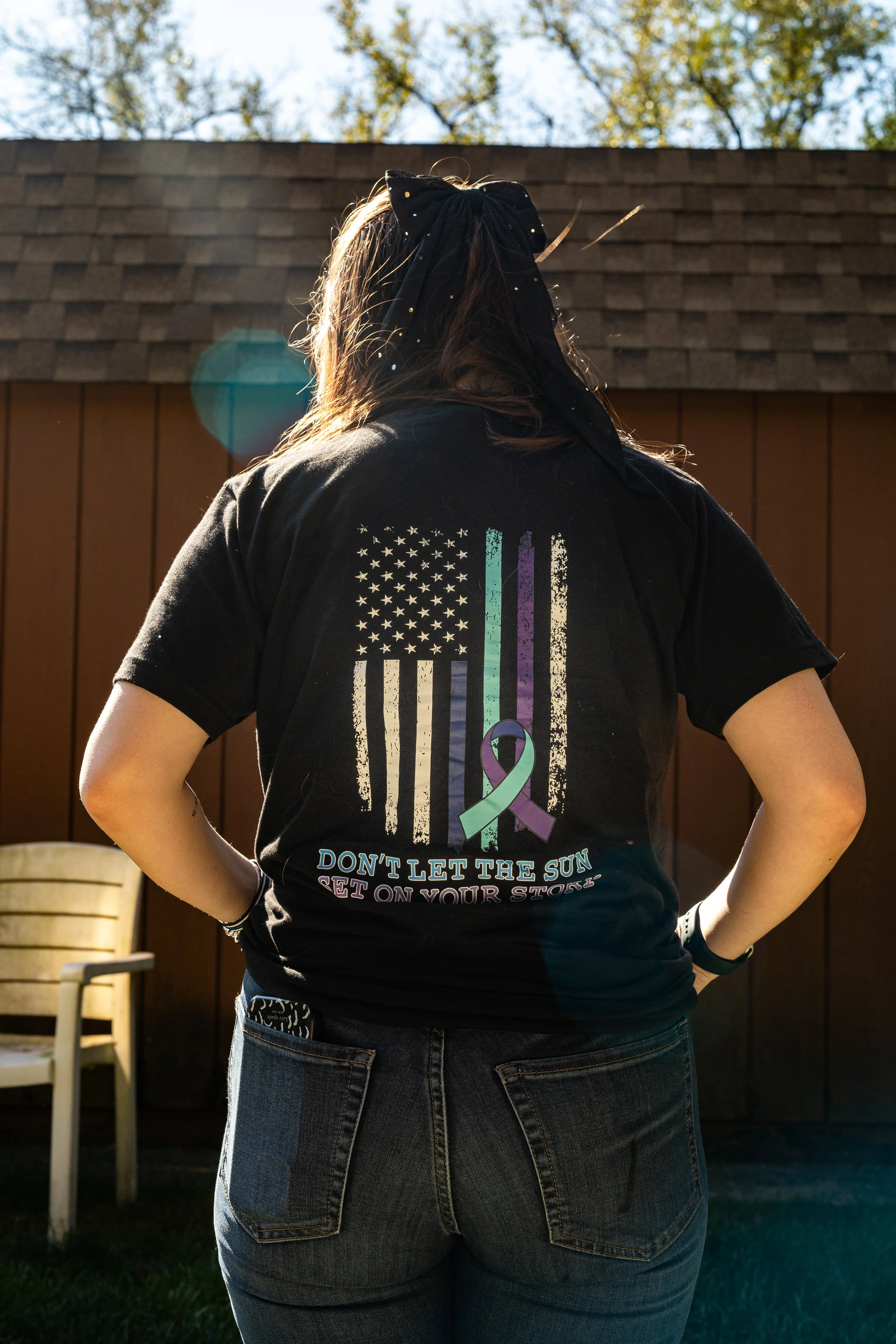Person wearing a black t-shirt with a design of a distressed American flag in black and white, overlayed with teal and purple stripes, and a purple awareness ribbon. The shirt has a message: 'Don't let the sun set on your story.' The person has long 
