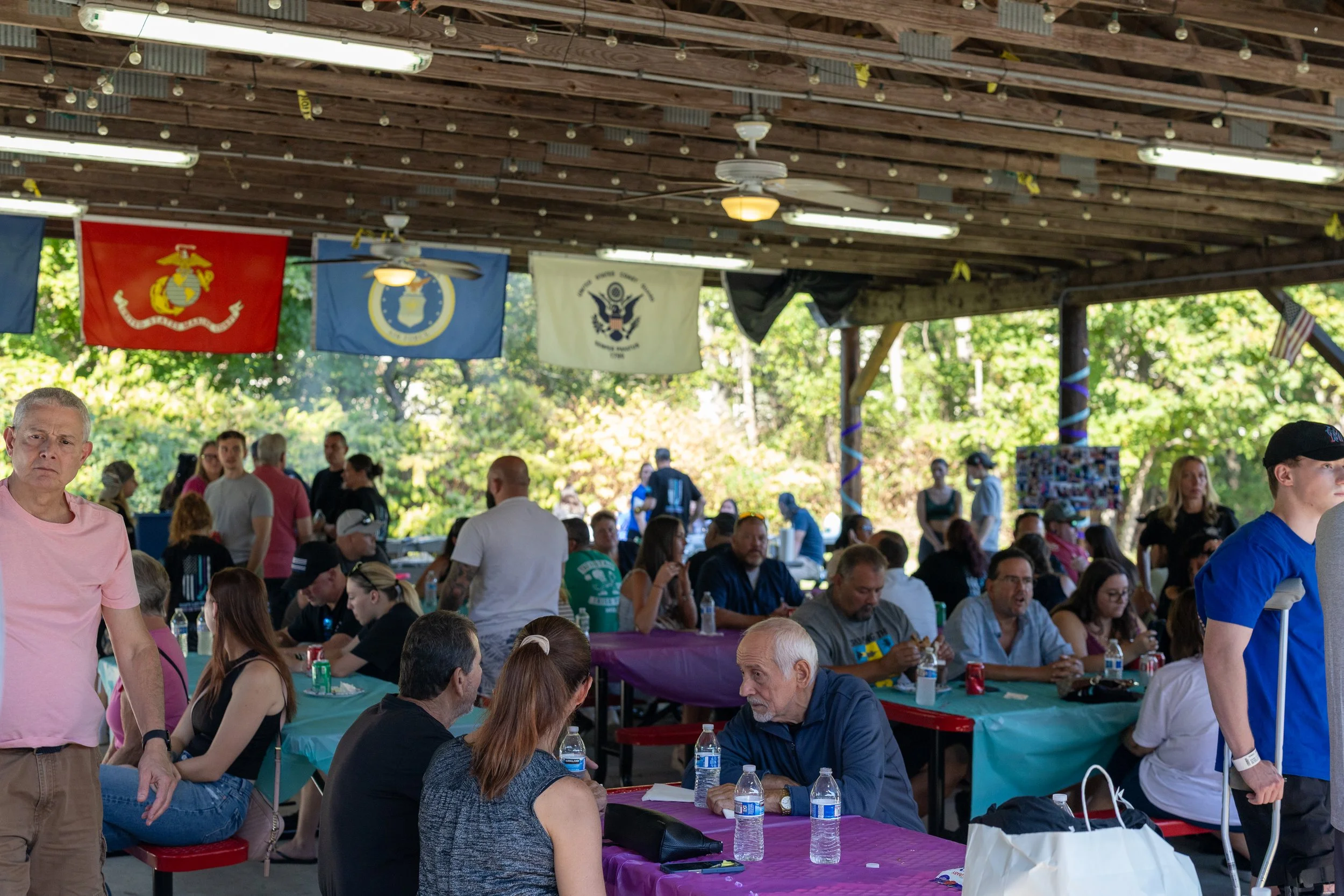 People gathered under a pavilion at an outdoor event, with military flags hanging from the ceiling and trees visible in the background.