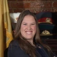A woman smiling in front of a brick wall with a yellow flag and a black hat with gold decorations.