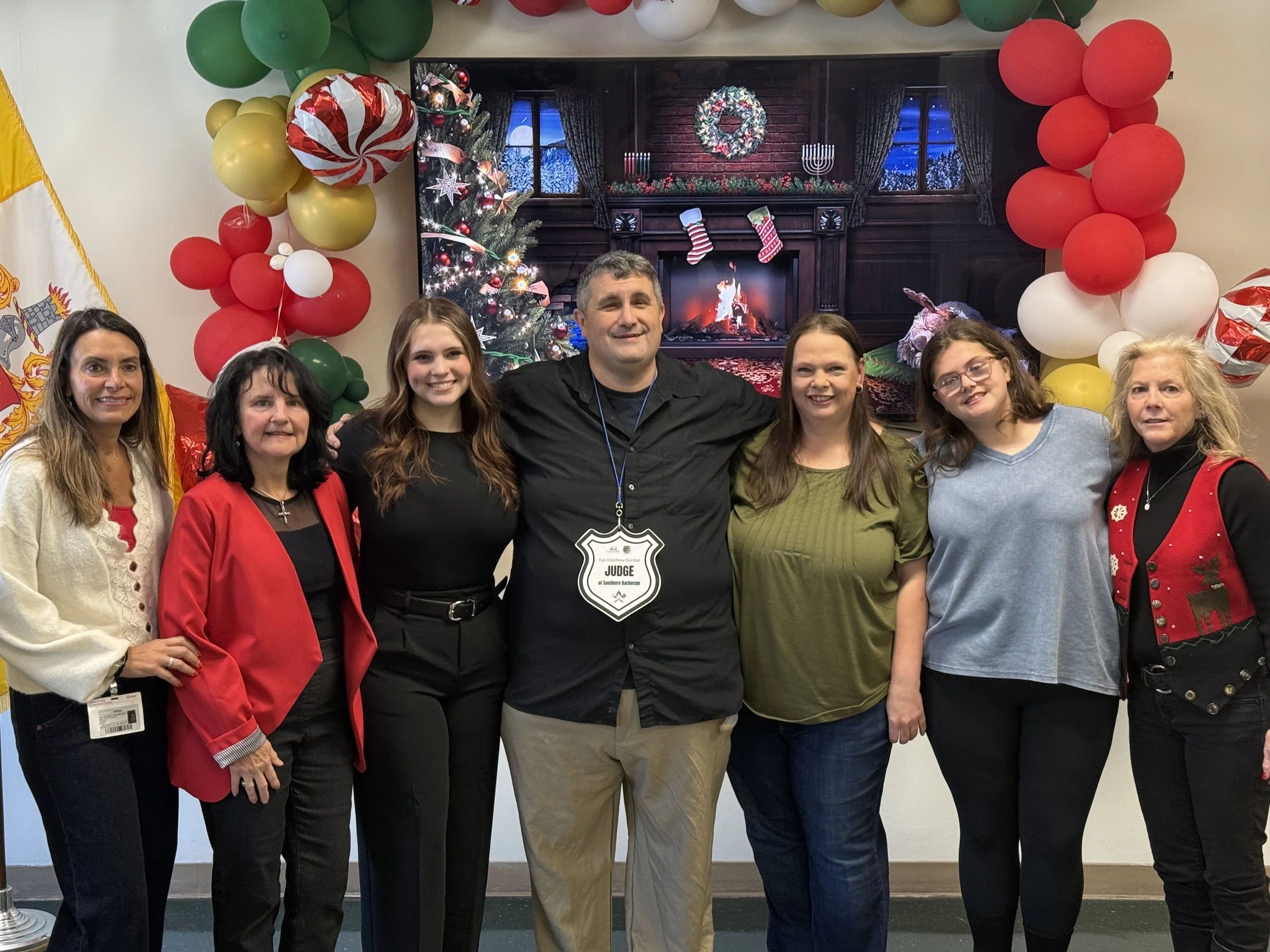 Group of seven people standing in front of a Christmas-themed background with red, green, white, and gold balloons, and a large screen displaying a decorated fireplace with Christmas stockings and a Christmas tree.