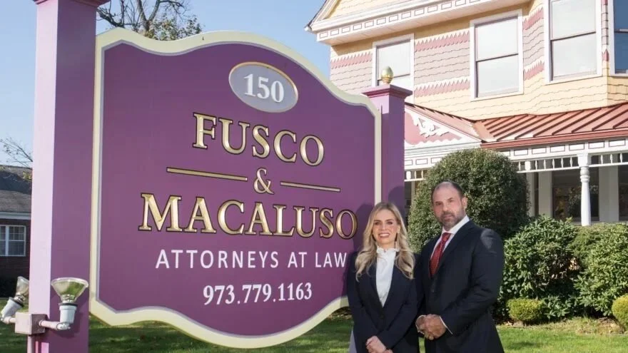 Two lawyers, a woman and a man, standing in front of a purple law office sign for Fusco & Macaluso Attorneys at Law, with a house and bushes in the background.