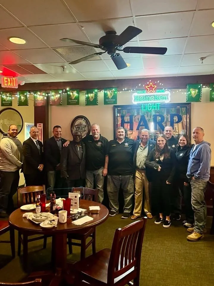 Group of people posing for a photo inside a restaurant or pub, with a table in the foreground and festive decorations including a neon sign, Guinness banners, and a wall art saying "HAPPY" in the background.