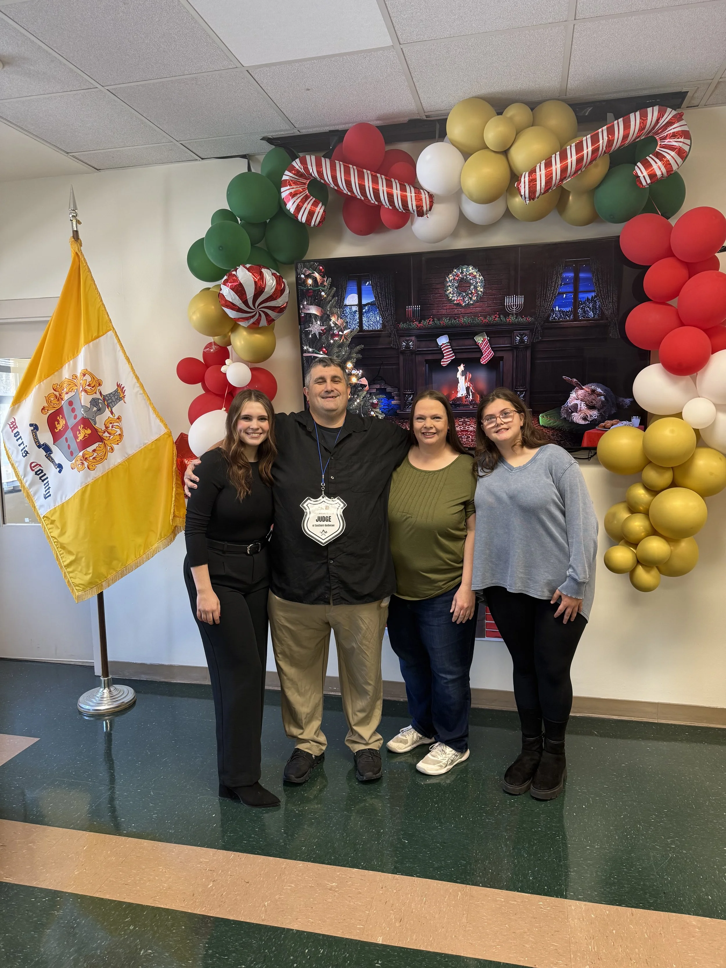 Group of four people standing in front of a holiday decorated background with balloons and a Christmas fireplace screen, and a flag on the left side.