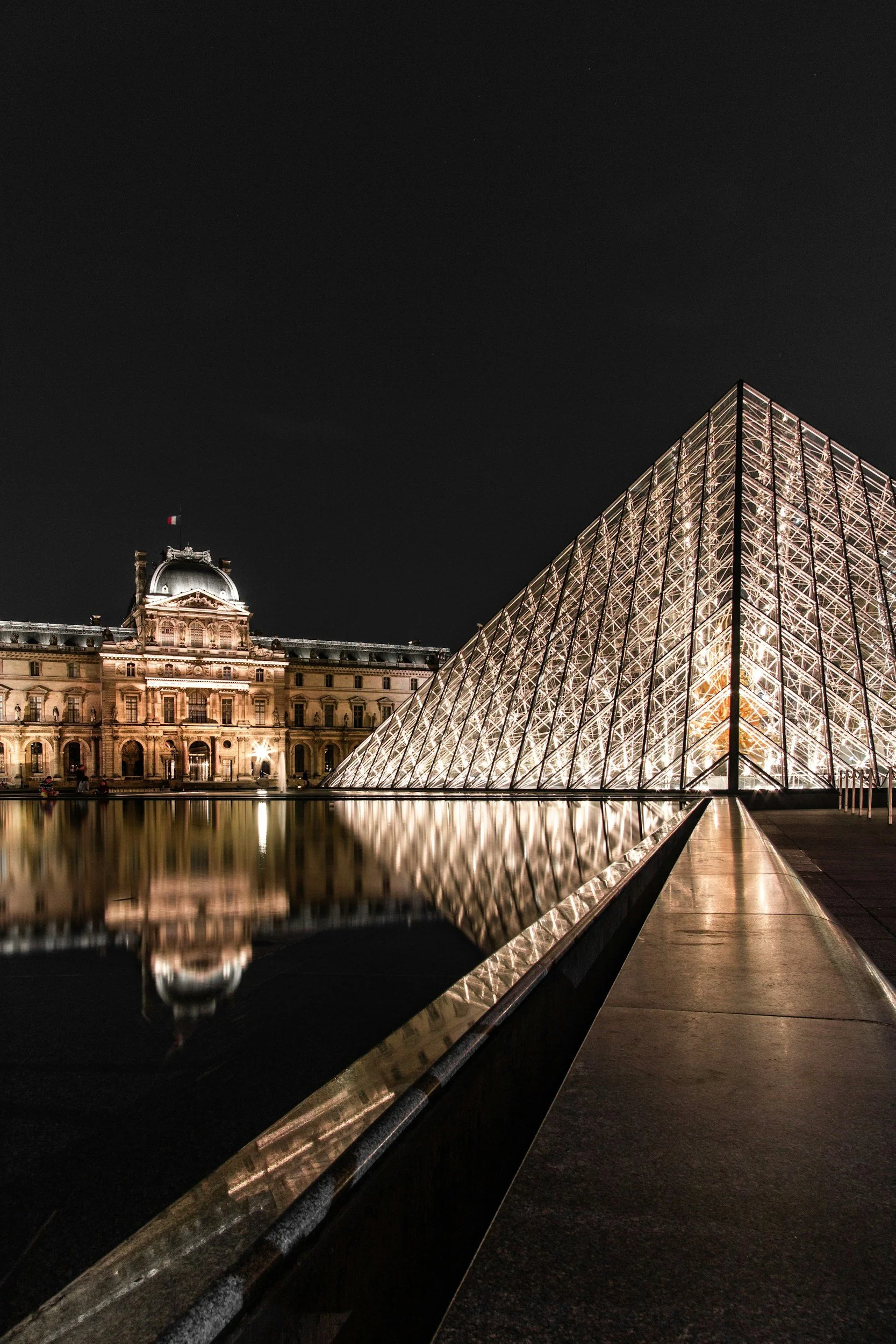 Night view of the Louvre Pyramid in Paris, France, with the historic Louvre Palace building in the background, reflected in a water feature.