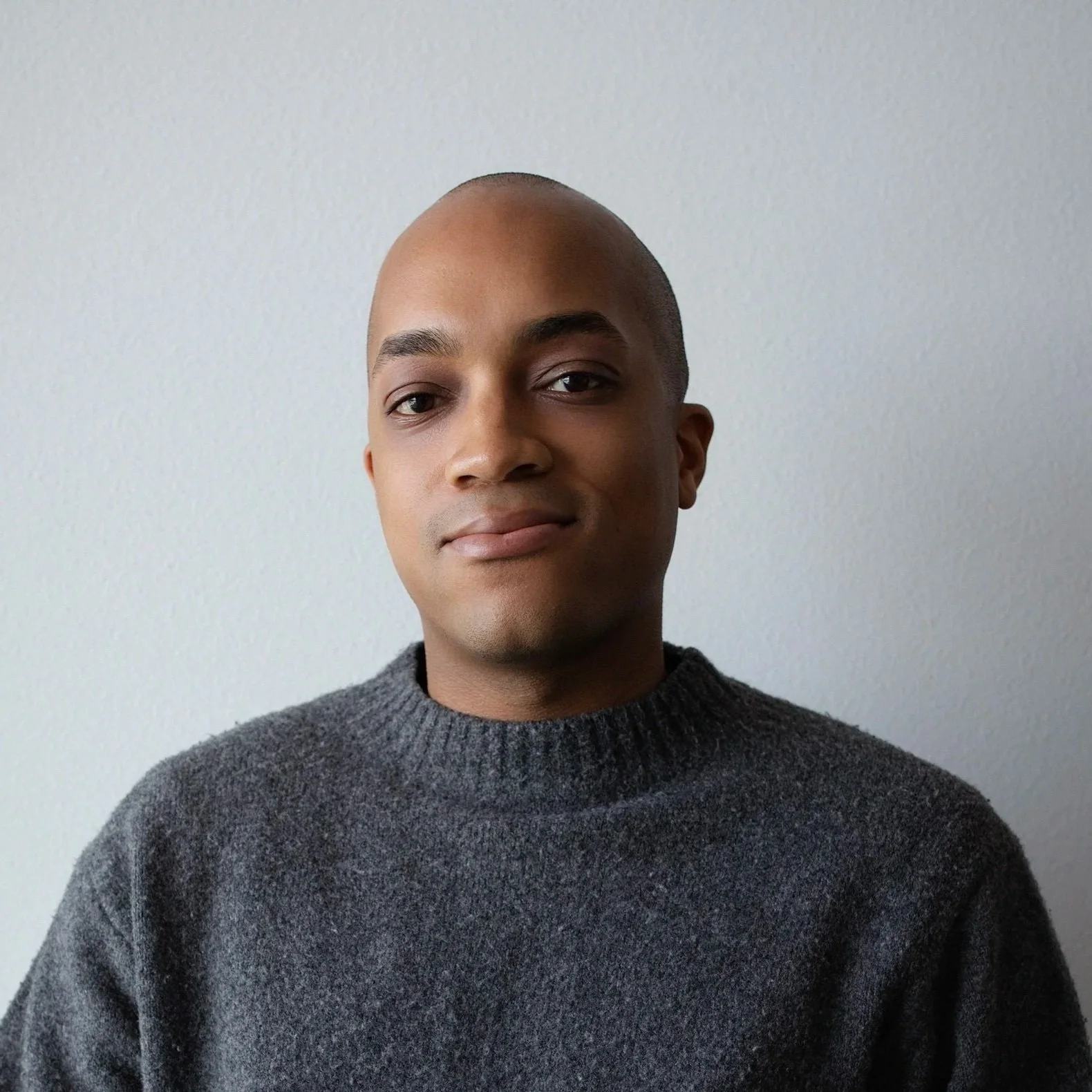 Portrait of a young man, wearing a dark gray sweater, standing against a plain light gray wall.