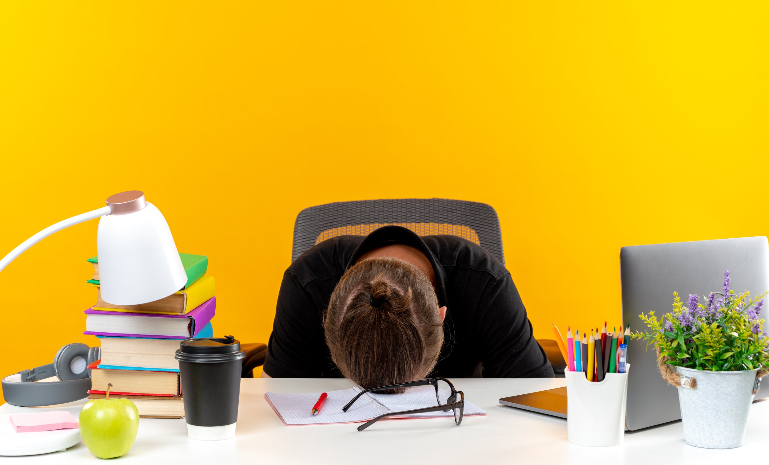 Person with hair in bun resting head on desk in an office, with yellow wall background, surrounded by office supplies and plants.