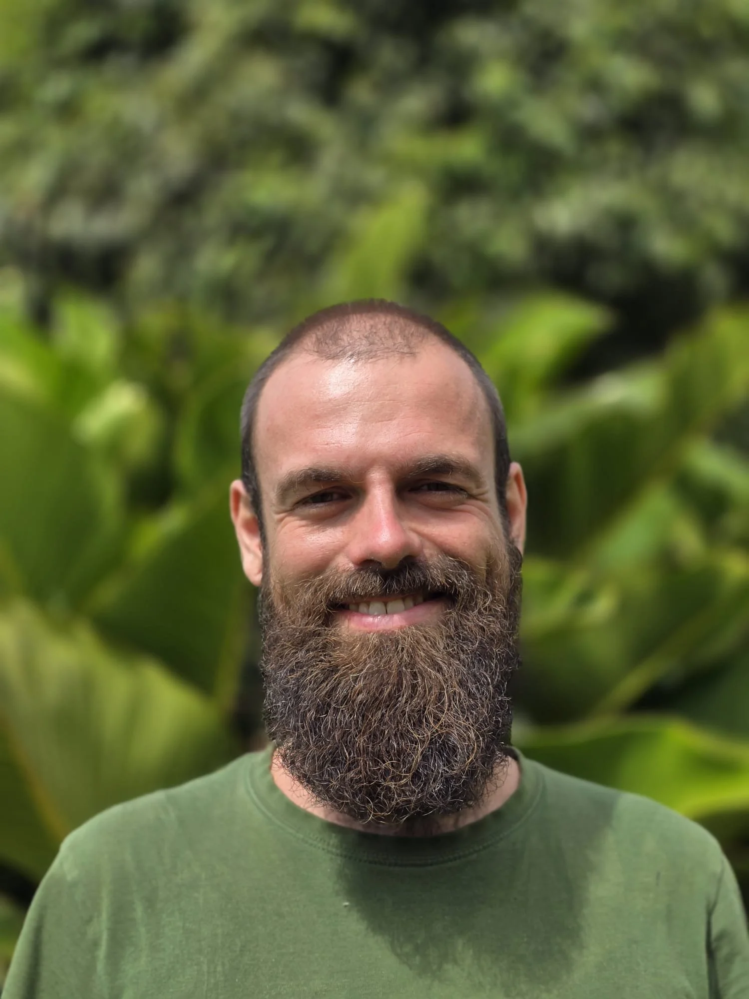 A smiling man with a full beard and short hair, wearing a green shirt, standing outdoors with lush green foliage in the background.
