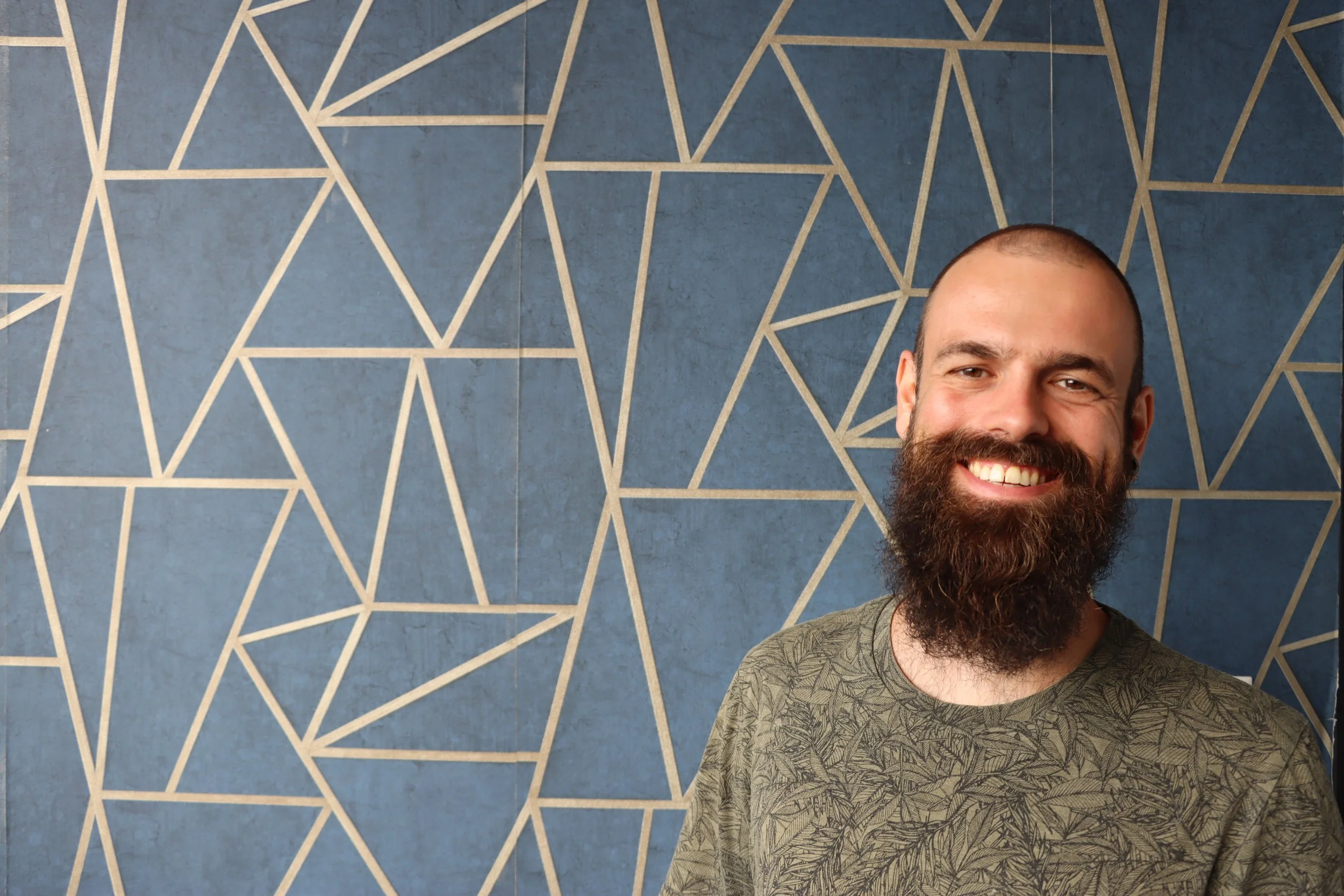 A man with a beard smiling, standing in front of a blue wall with a geometric white line pattern.
