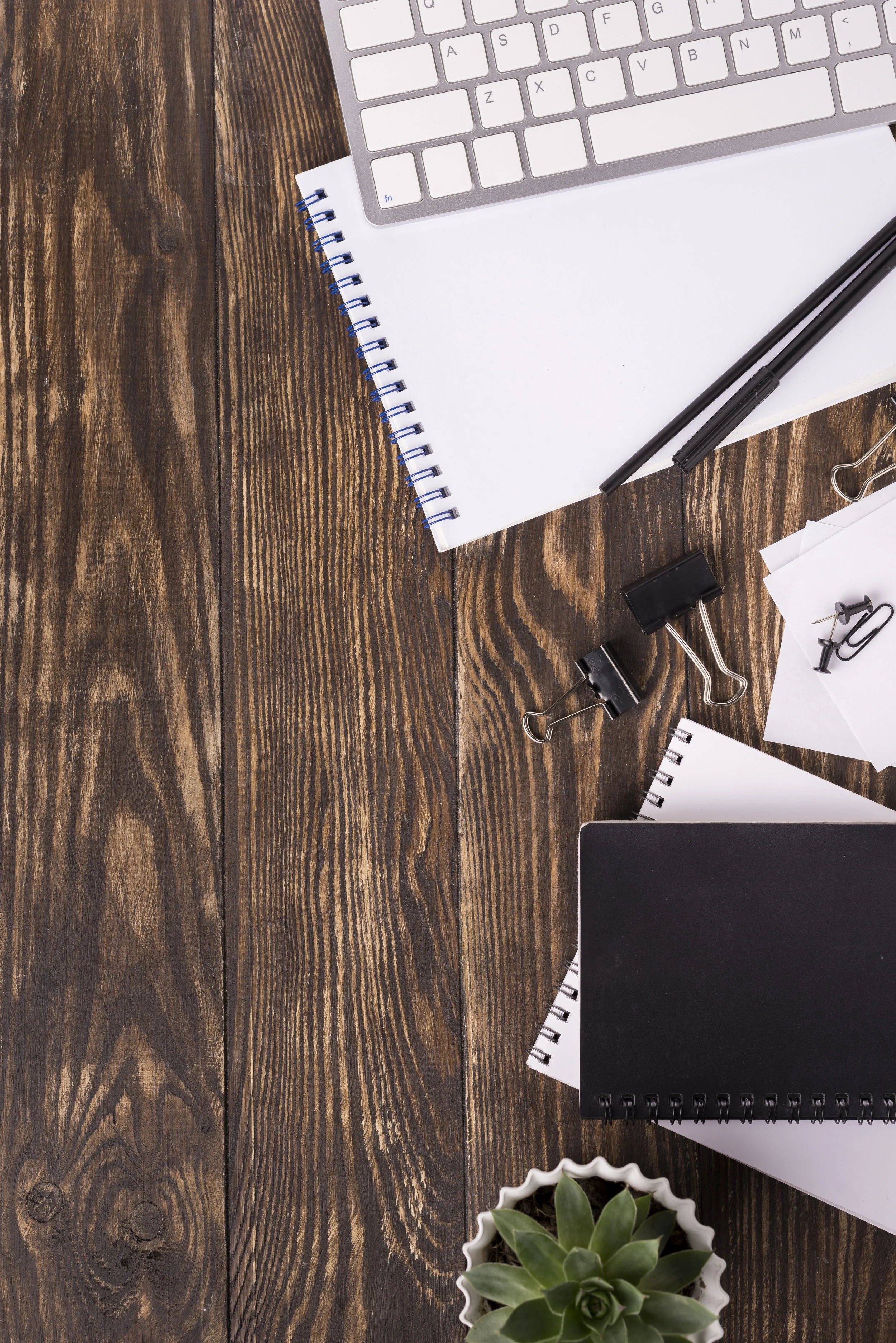 Top view of a wooden desk with office supplies, including a white keyboard, spiral notebooks, black pen, binder clips, sheets of paper, a black notebook, and a small potted succulent plant.
