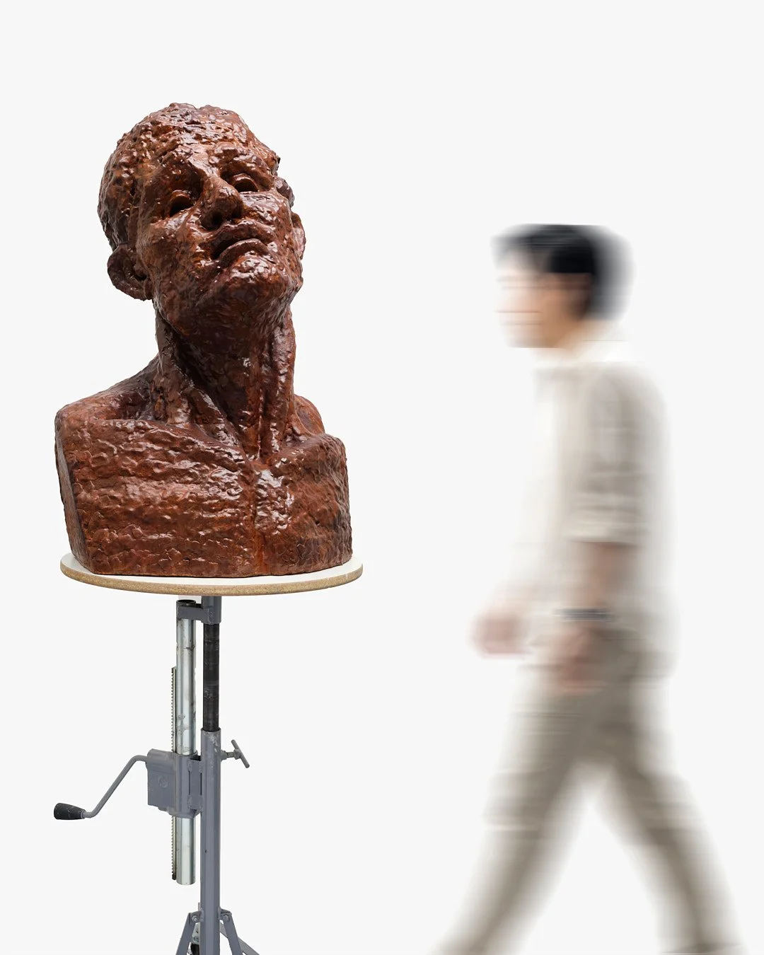 A bronze bust sculpture of a man's head and shoulders on a stand, with a blurred person walking past in the background.