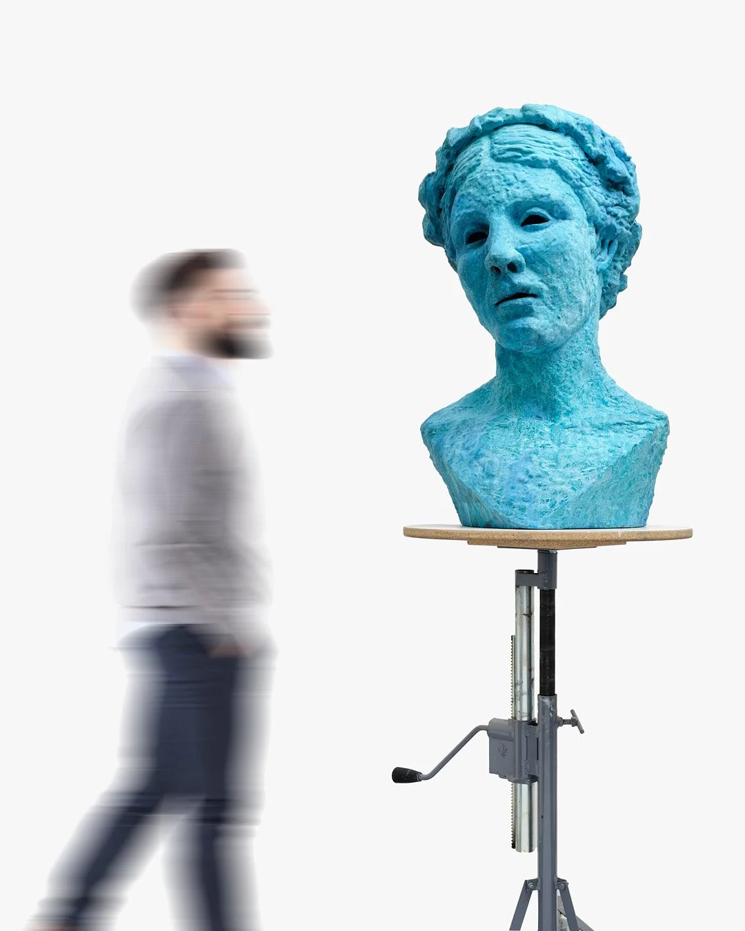 A man with a beard in a gray sweater stands near a blue sculpture of a woman's bust on a stand against a white background.