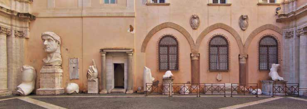 Exterior view of a historical building with several white marble sculptures and busts, including a large head sculpture on a pedestal, set against a peach-colored wall with arched windows.