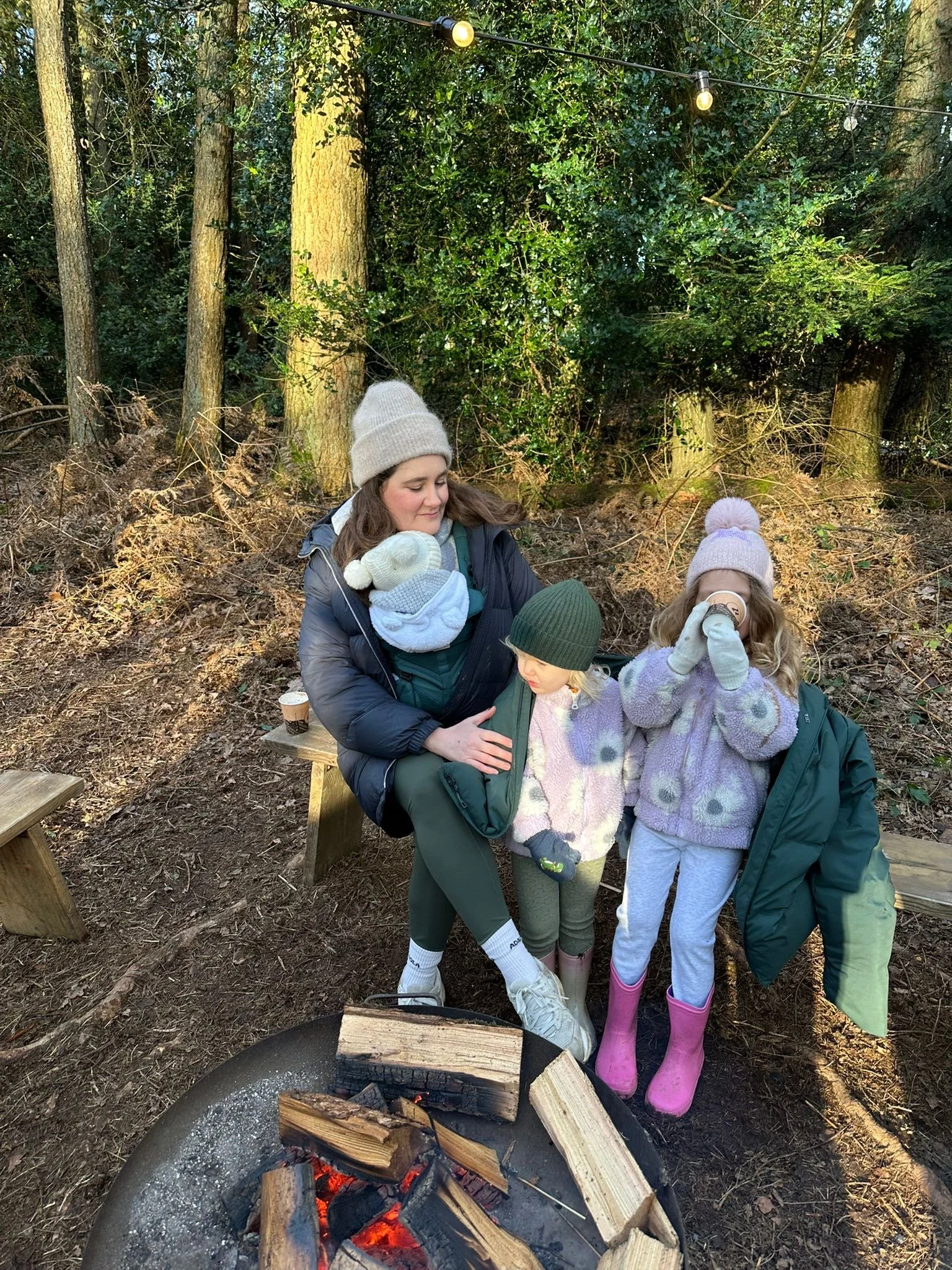 A woman with two young girls outdoors around a campfire in a wooded area, wearing jackets, knit hats, and boots, with string lights overhead.