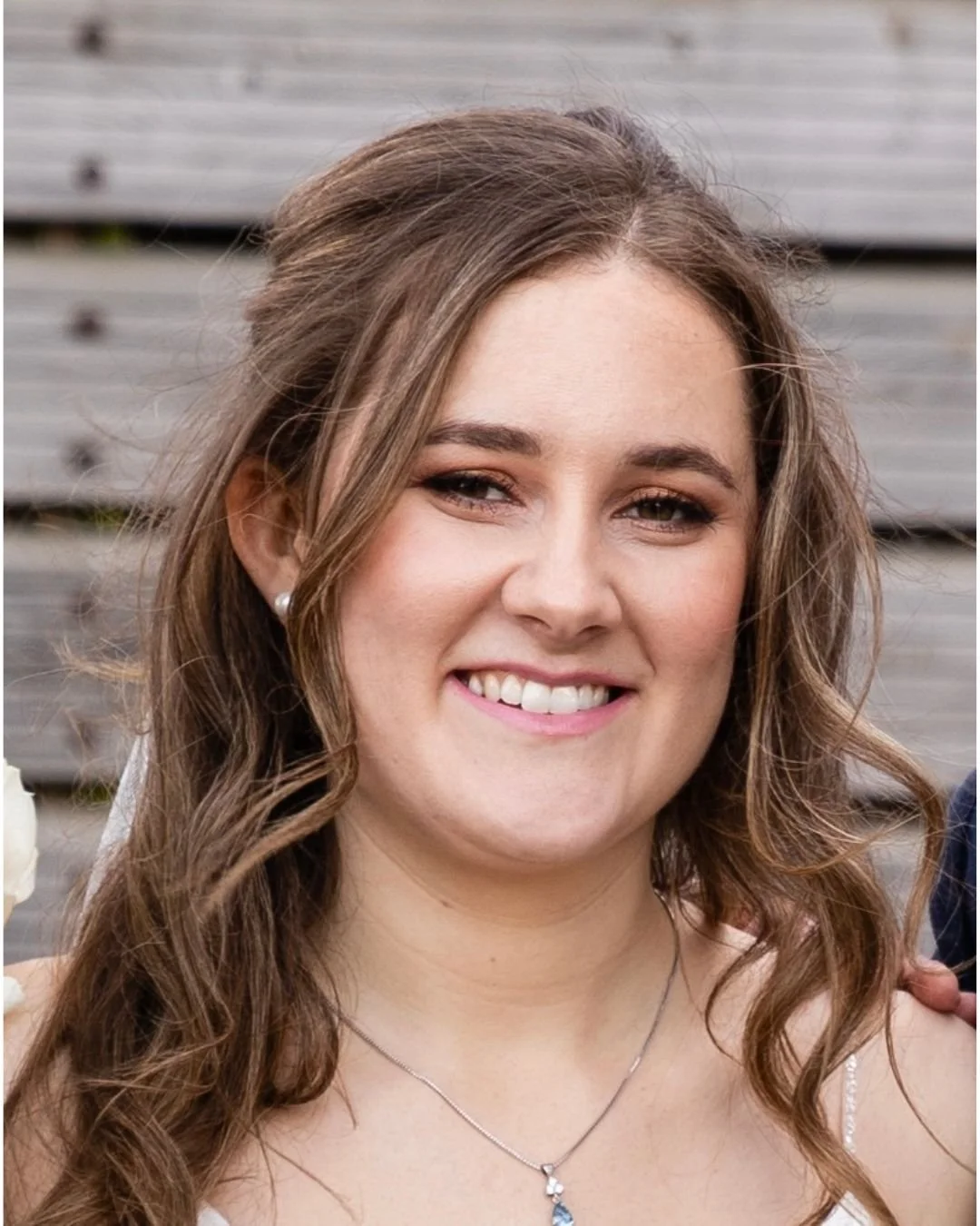 Close-up of a smiling woman with long wavy brown hair, wearing pearl earrings and a necklace, standing outdoors with a wooden fence background.