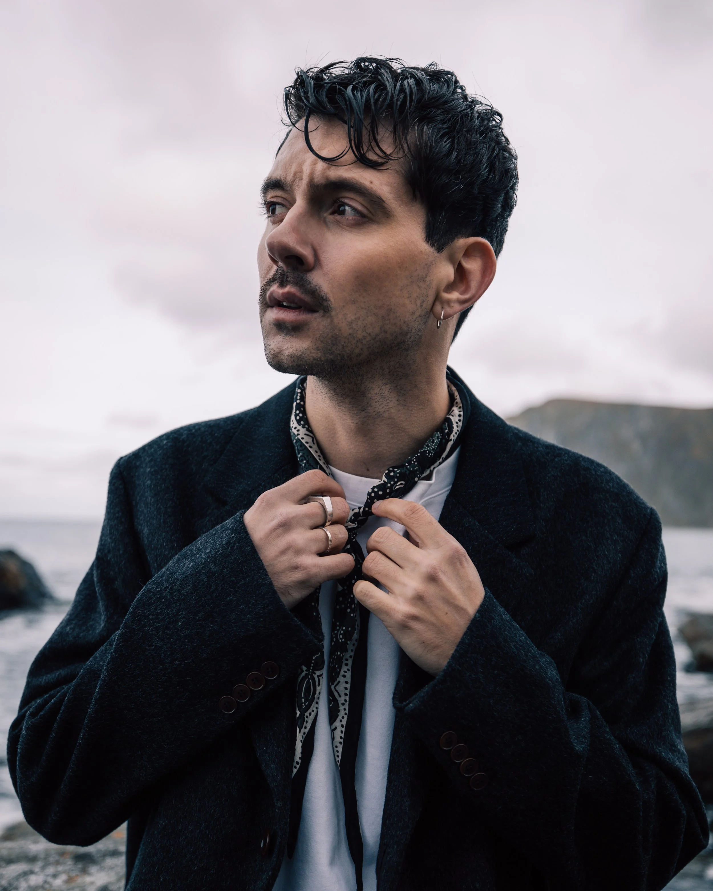A man with dark curly hair, facial hair, and an earring on his left ear, looking to his left. He's adjusting a black and white polka-dotted scarf or tie around his neck, wearing a dark coat, set against a cloudy, overcast sky with a blurred coastal background.
