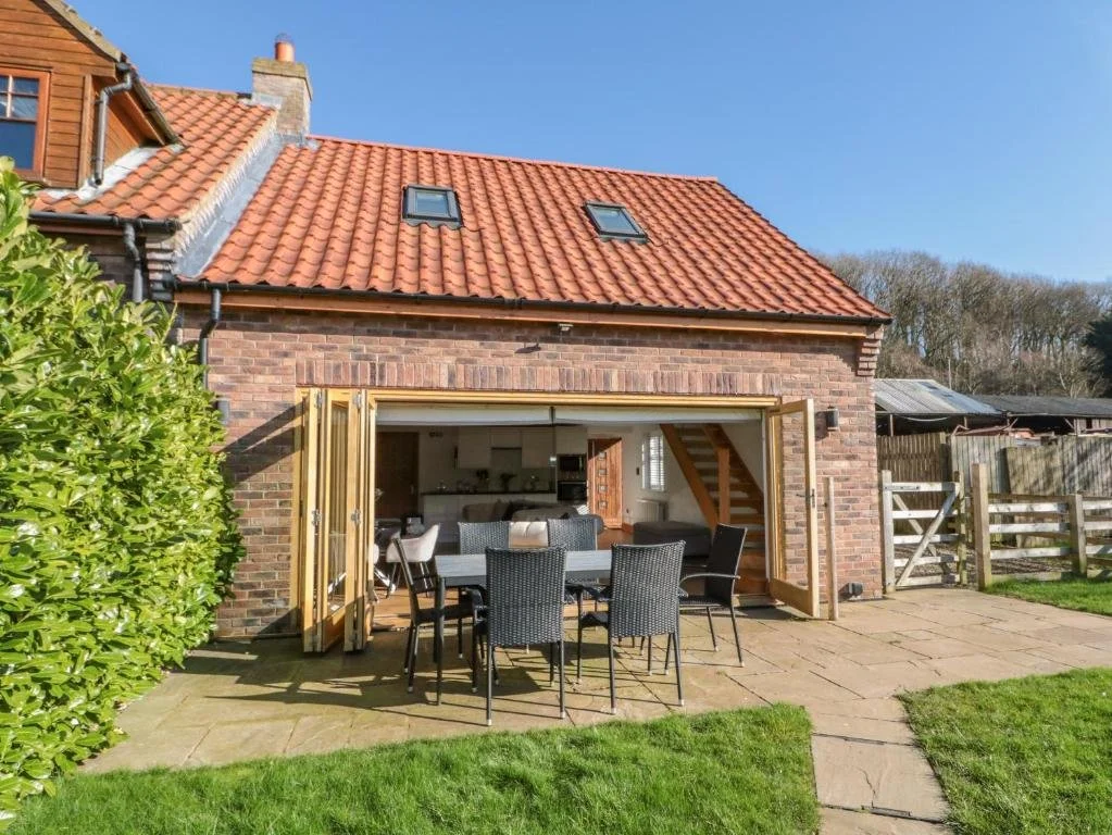 Backyard with a brick house and an outdoor dining area with a table and six chairs, open doors revealing a kitchen inside, and a fenced yard with greenery.