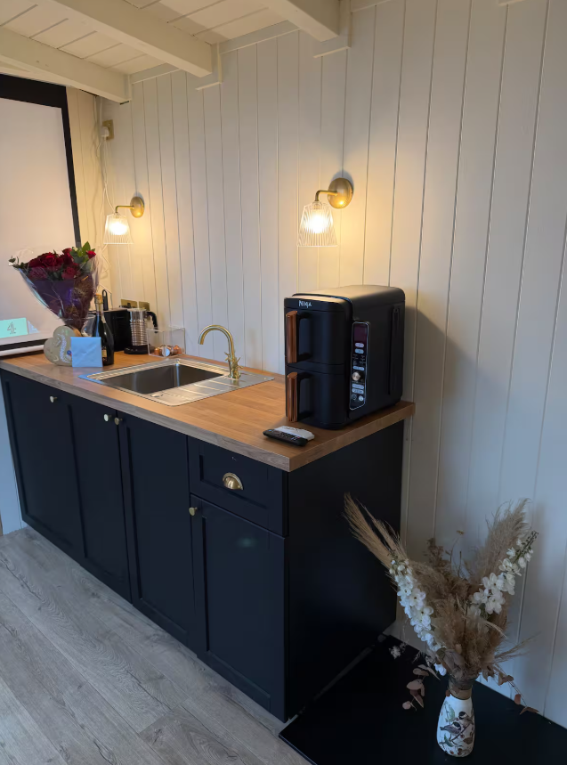 Kitchen area with a black coffee maker, a small sink with a gold faucet, and a remote on a wooden countertop. Wall-mounted lights above and a vase with dried flowers on a small black table beside it.