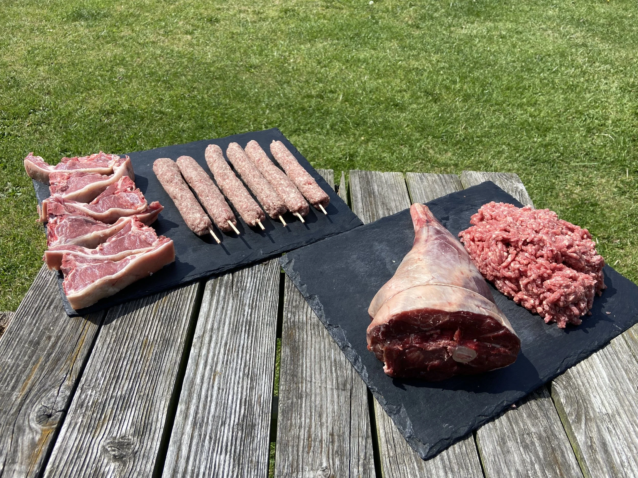 Raw lamb chops, skewered ground lamb kebabs, a lamb shoulder, and ground beef on black slate boards outdoors on a wooden table.