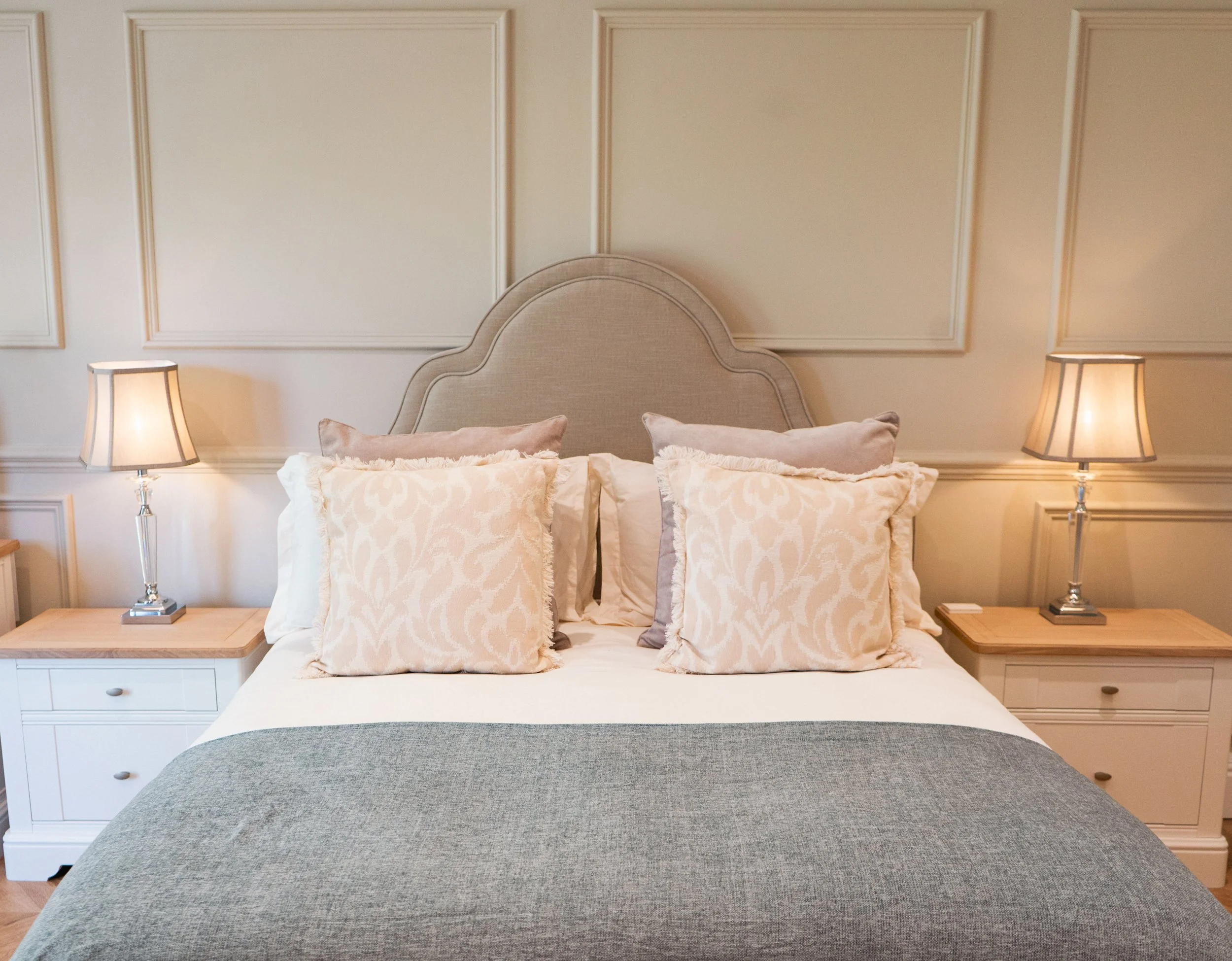 A neatly made bed with a beige upholstered headboard, multiple decorative pillows, and a gray and white bedding set, flanked by white nightstands with lamps.